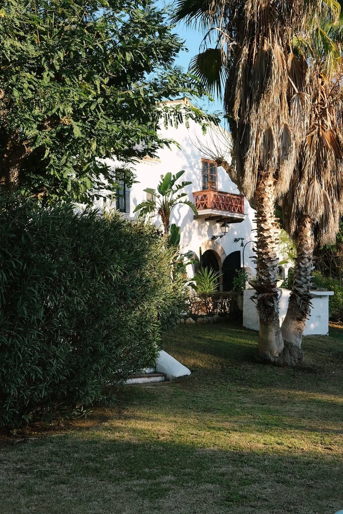 A white Spanish-style house with arched doorways, surrounded by lush green plants and tall palm trees, with a small balcony and sunny blue sky.