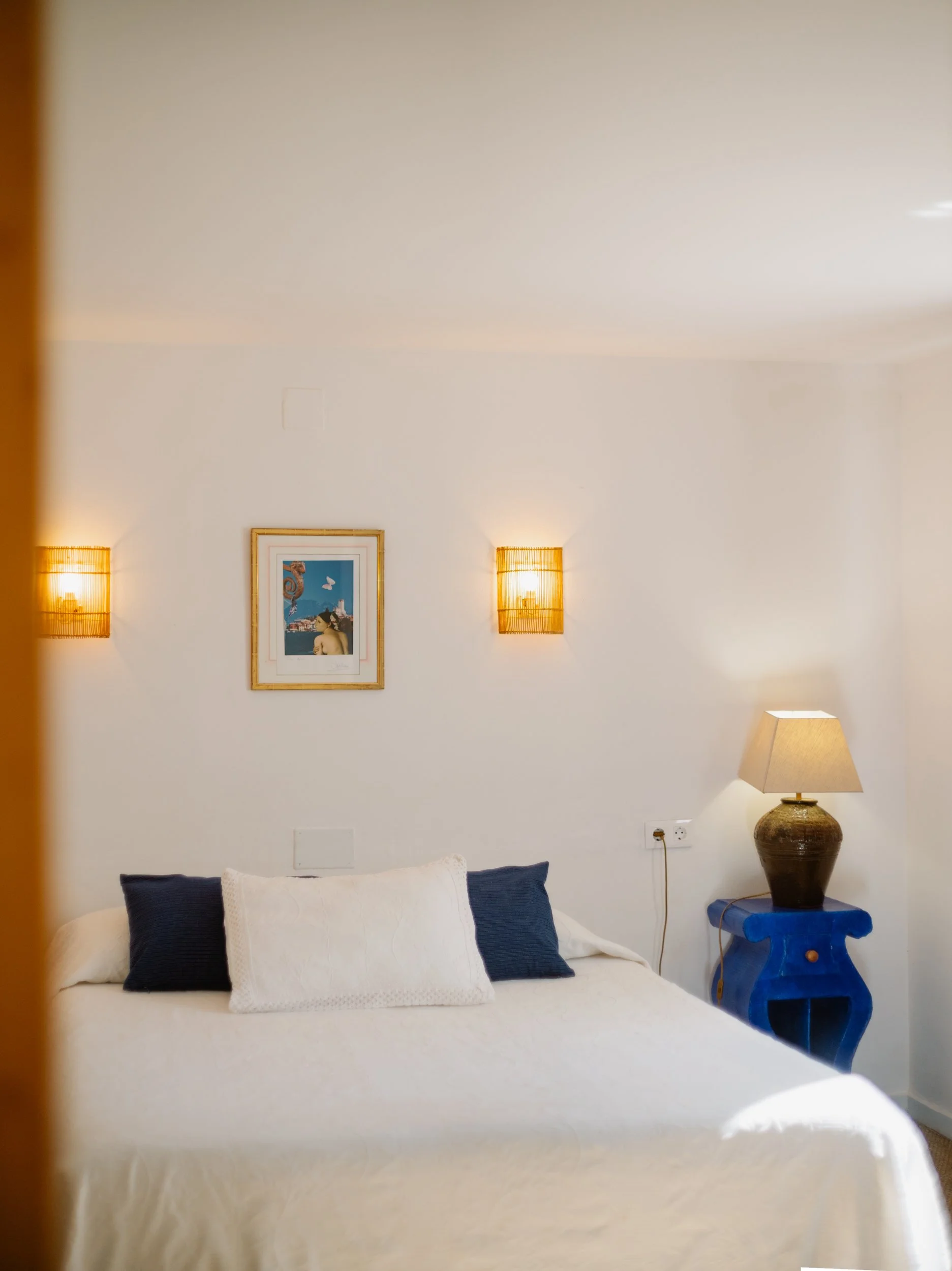 Chambre avec lit blanc, deux coussins bleus, une lampe de table et des appliques murales