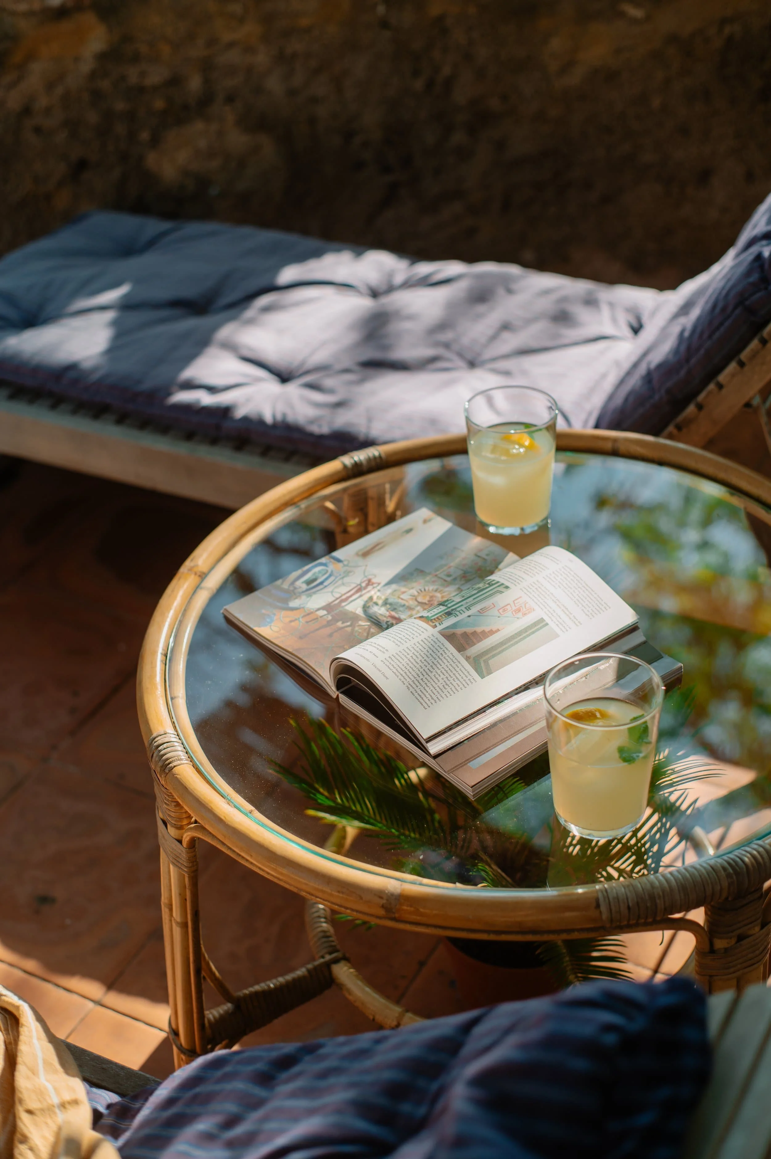 A round wicker table with a glass top holds an open magazine and two glasses of lemonade with lemon slices. There is a lounge chair with a cushion in the background.
