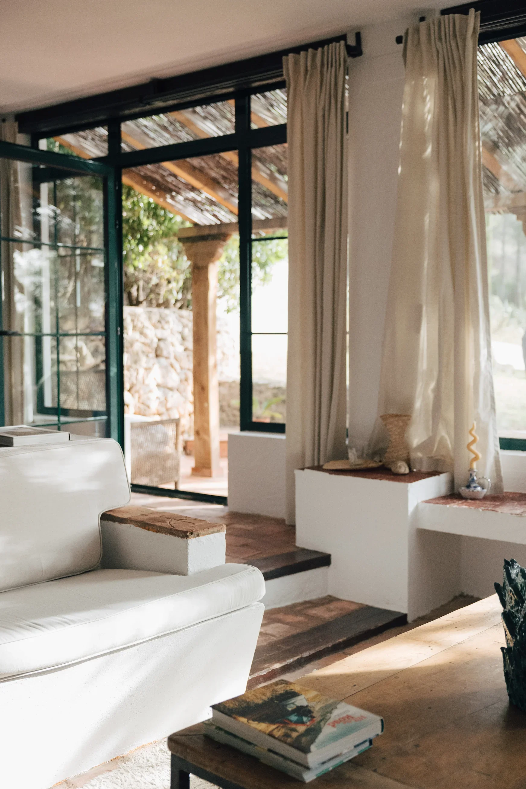 Intérieur d'un salon lumineux avec un canapé blanc, une table en bois, et des rideaux crèmes devant de grandes fenêtres donnant sur une terrasse en plein air.