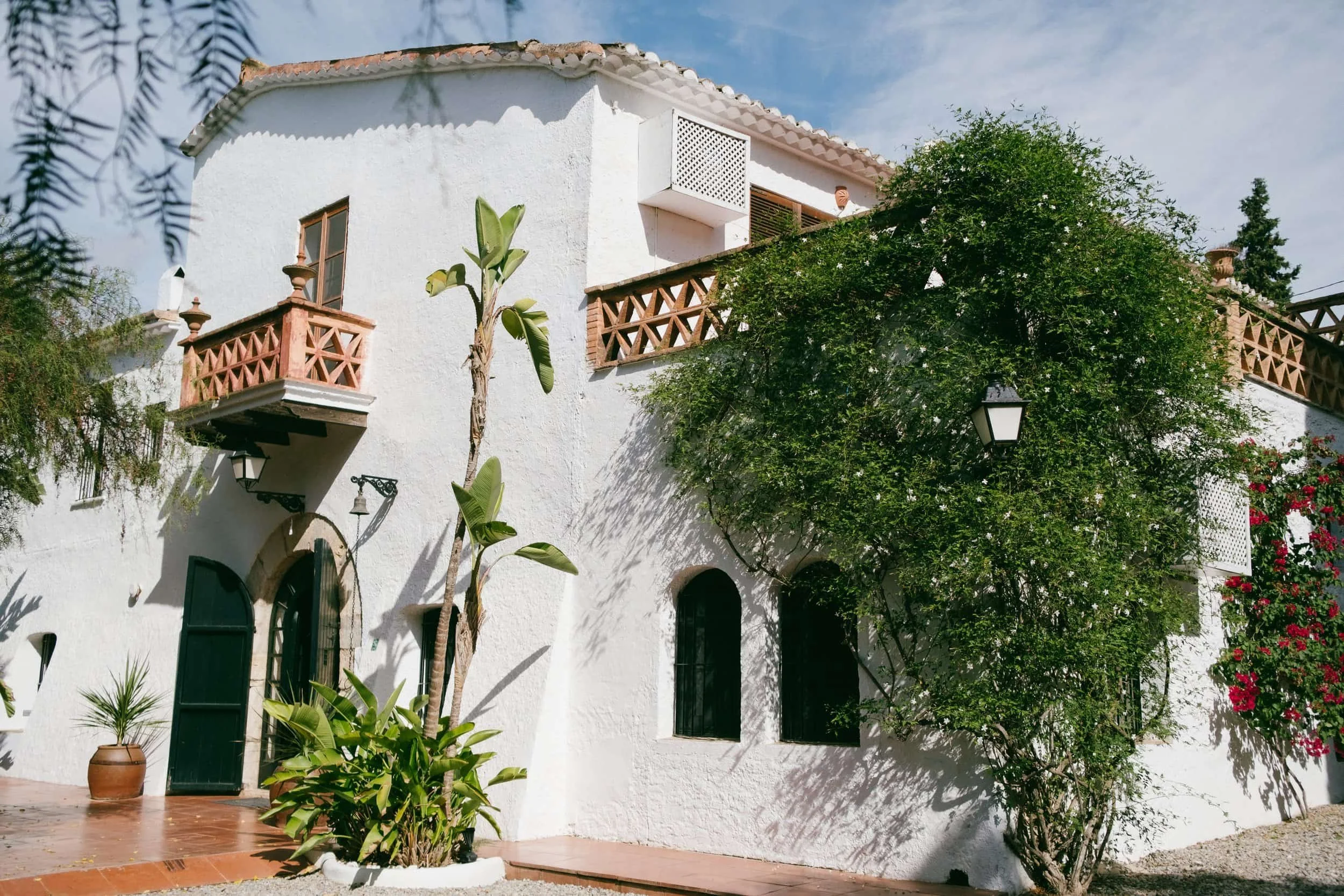 White Mediterranean-style house with terracotta roof, wooden balconies, arched windows, lush greenery, and potted plants outside.