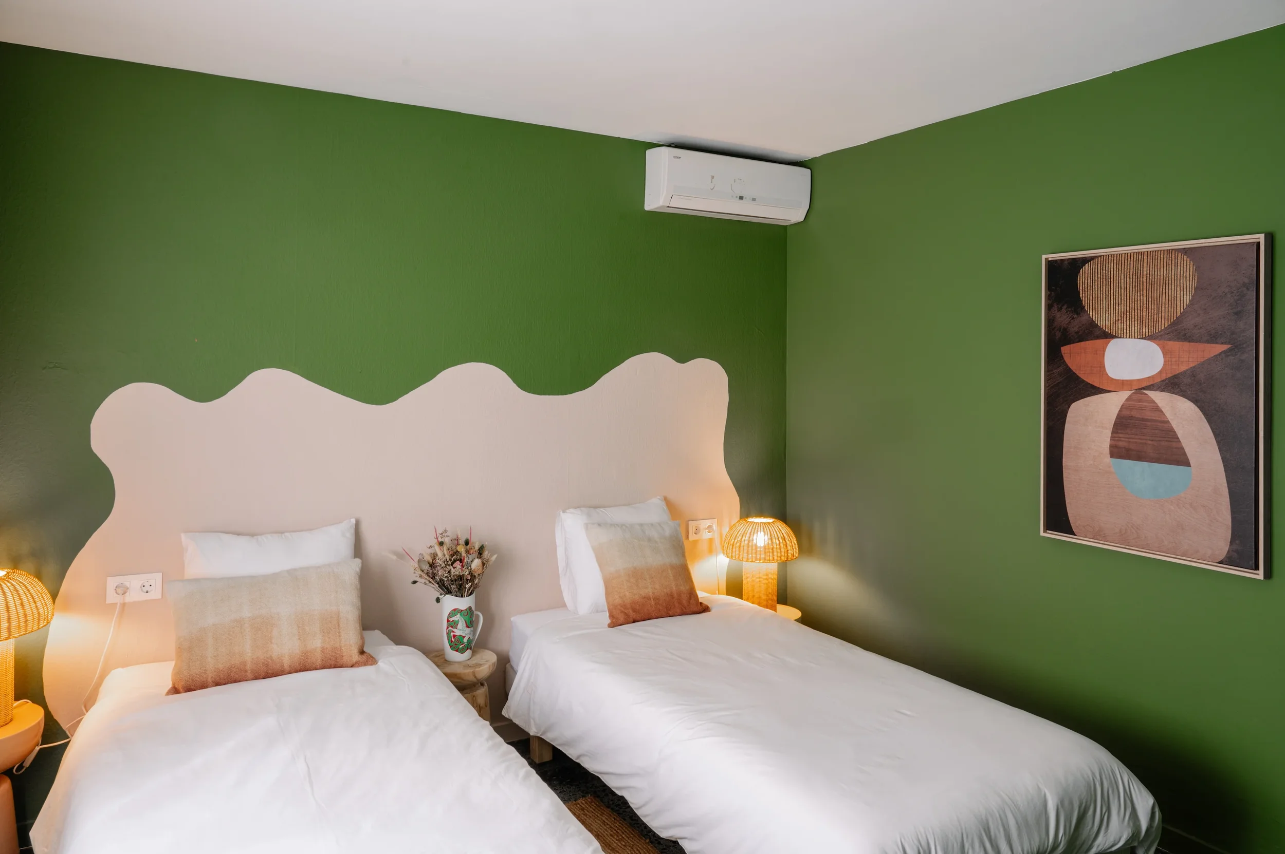 Chambre avec deux lits, tête de lit blanche, mur vert, lampe en osier, tableau abstrait, plante