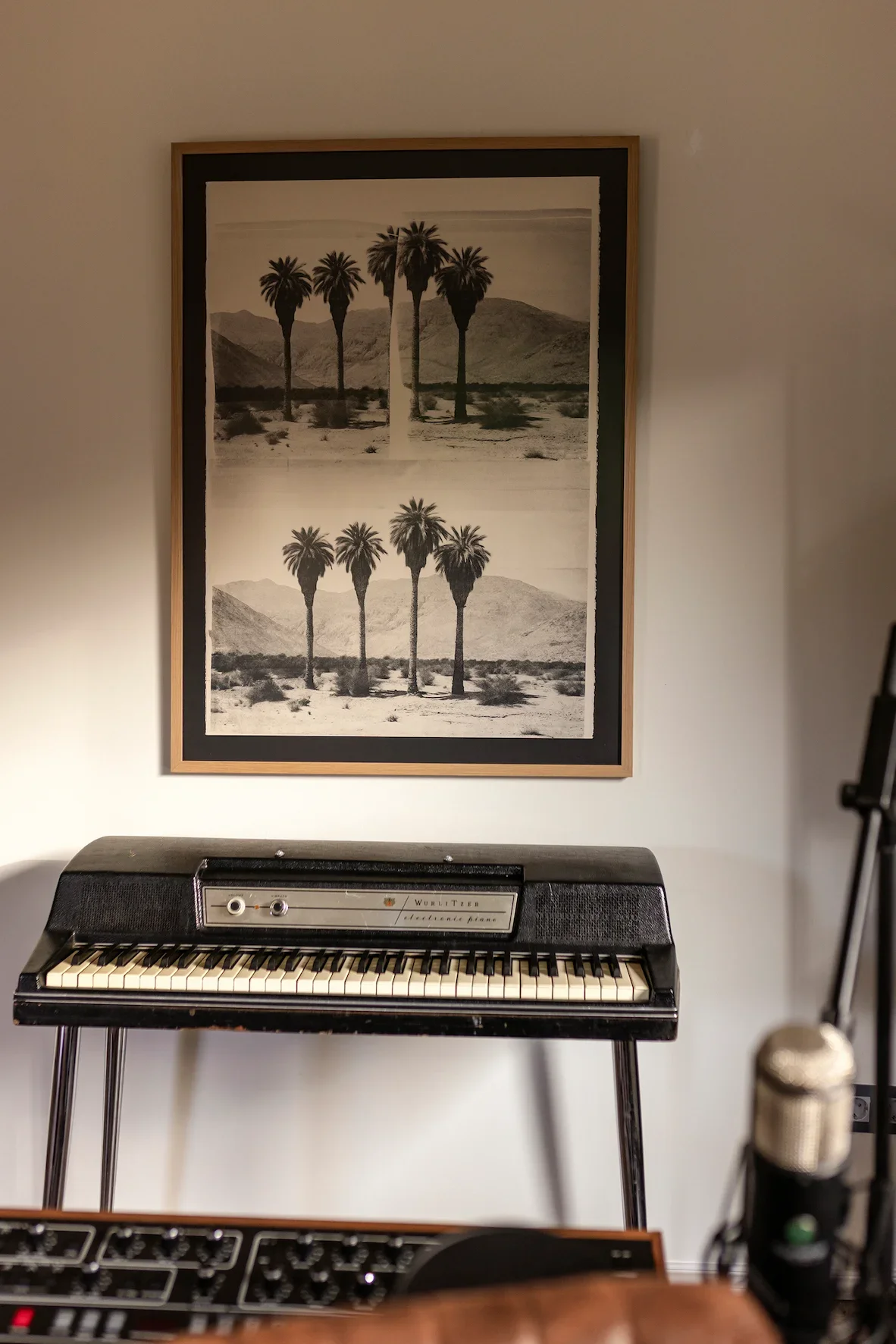 Une affiche monochrome représentant un paysage de palmiers et montagnes, avec deux rangées de palmiers, dans un style art contemporain, suspendue sur un mur blanc à côté d'un clavier électrique vintage sur un support.