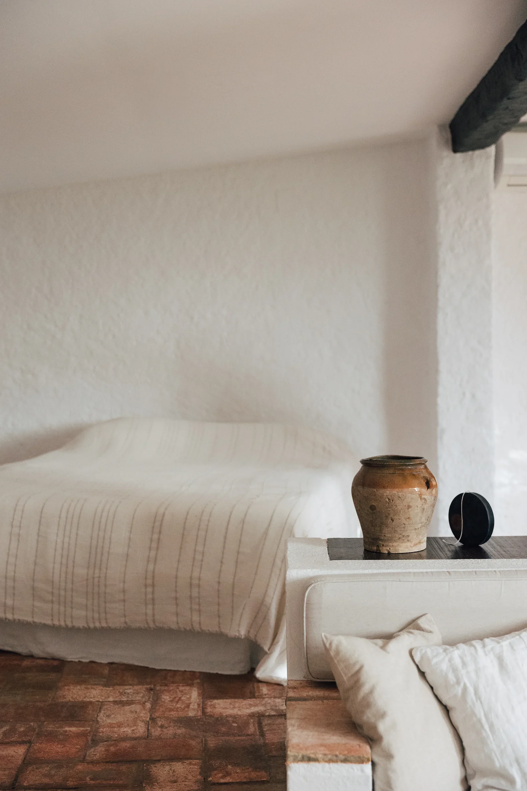 Intérieur d'une pièce avec mur blanc, lit avec drap beige, pot en terre cuite et petite enceinte noire sur une table blanche en bois, sol en briques rouges, coussins blancs au premier plan.