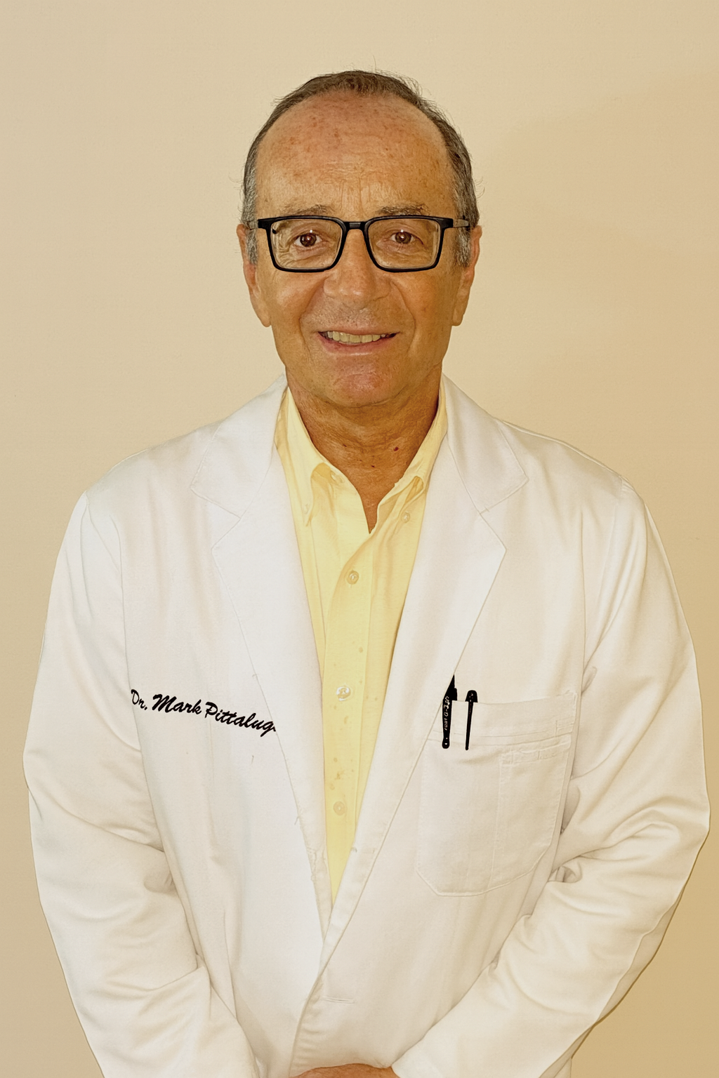 A smiling man in a white lab coat standing in front of framed diplomas or certificates on a wall.