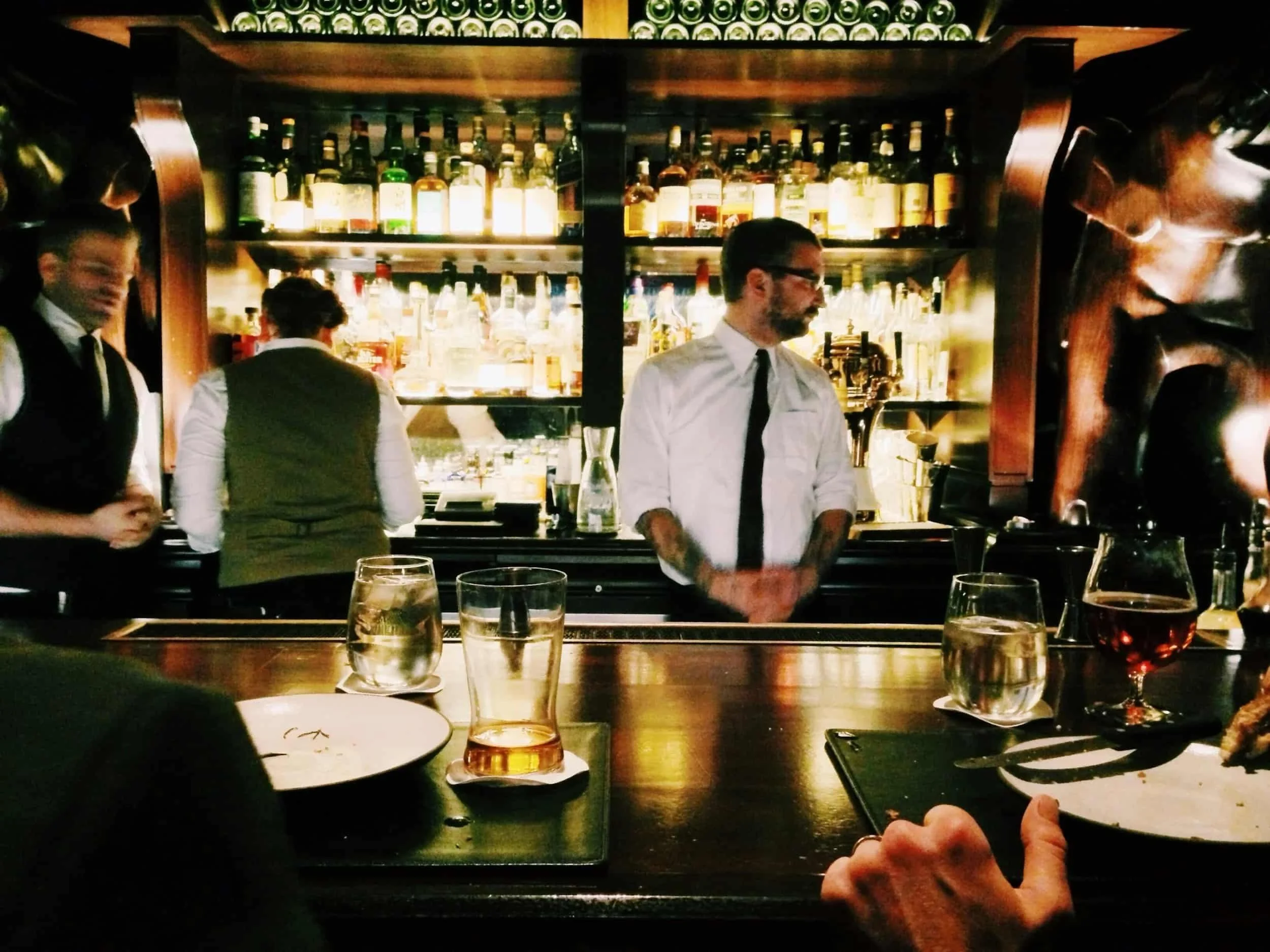 Bartenders working behind a dimly lit bar counter with bottles of liquor on shelves and drinks on the counter.