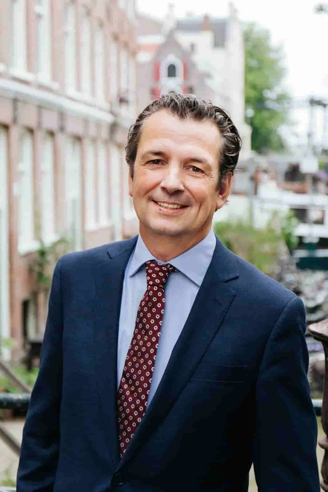 Gertjan Teeuwen glimlacht in een stadswijk, draag een donkerblauwe jas, lichtbl Forem en een donkerrode das met witte patronen, achter een ruit.