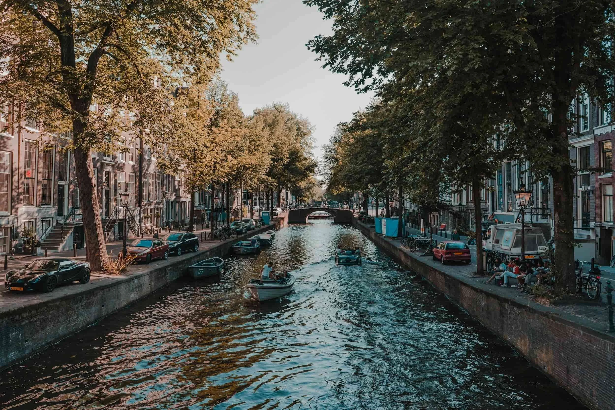 Een gracht in Amsterdam met wandelende mensen, boten op het water, en historische gebouwen langs de oever, omgeven door groene bomen.