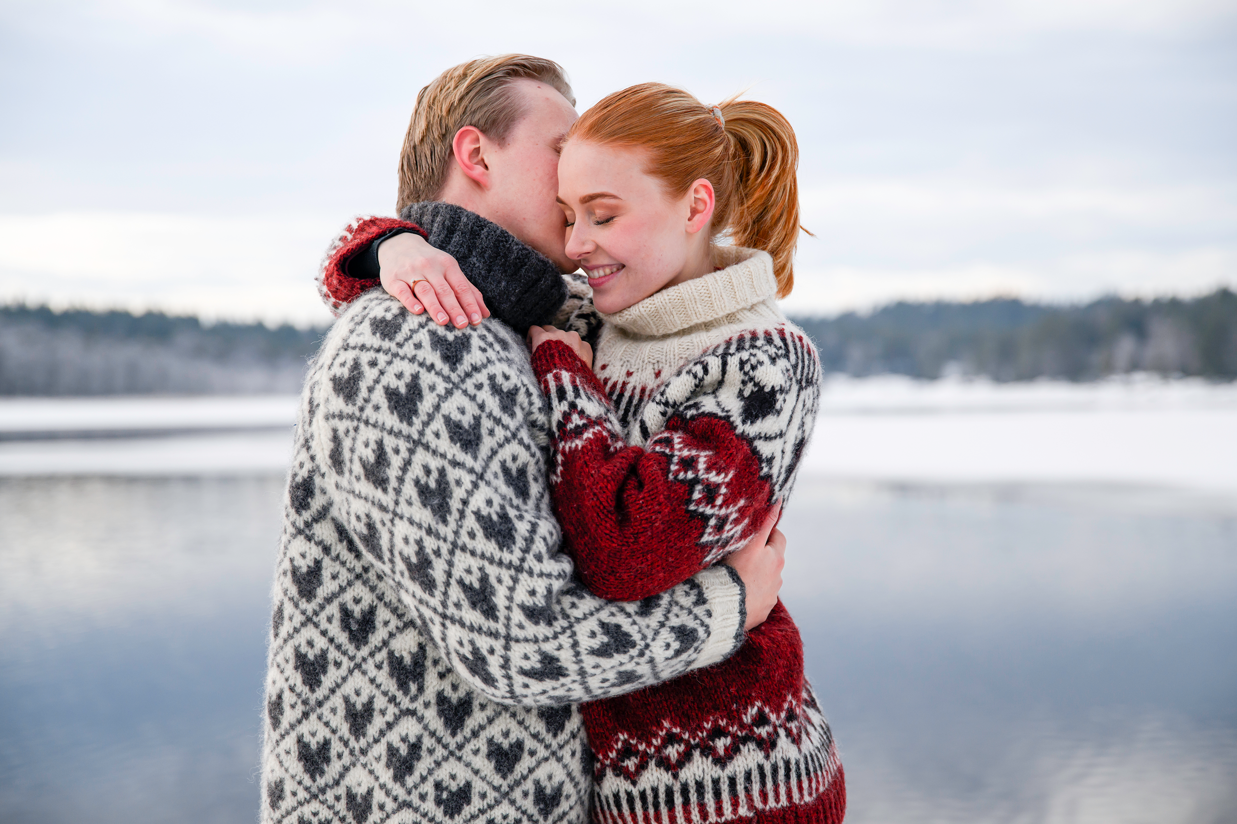 To personer omfavner ved en innsjø i kaldt vær, båret av vinterklær med mønstre. Utendørs vinterlandskap med vann og snø i bakgrunnen.