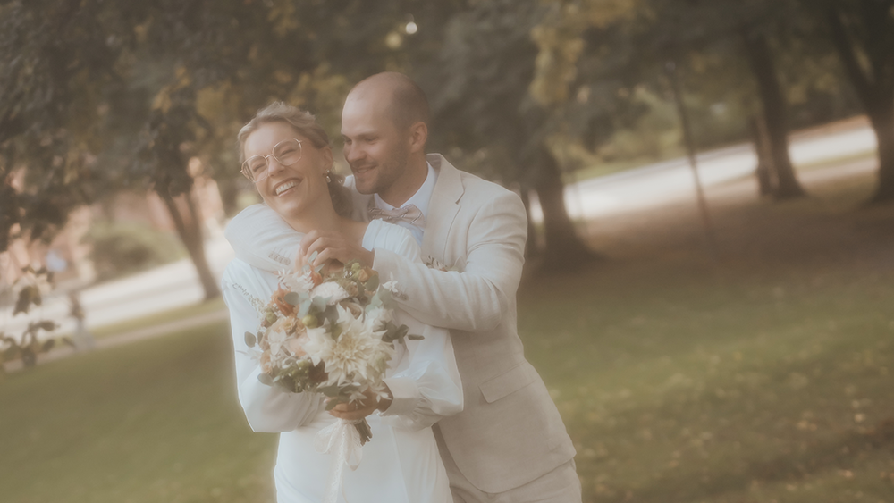 Mann og kvinne som er nygifte og smiler og ler fordi de har bryllup. De klemmer varmt hverandre mens kona holder bryllupsbuketten til seg.