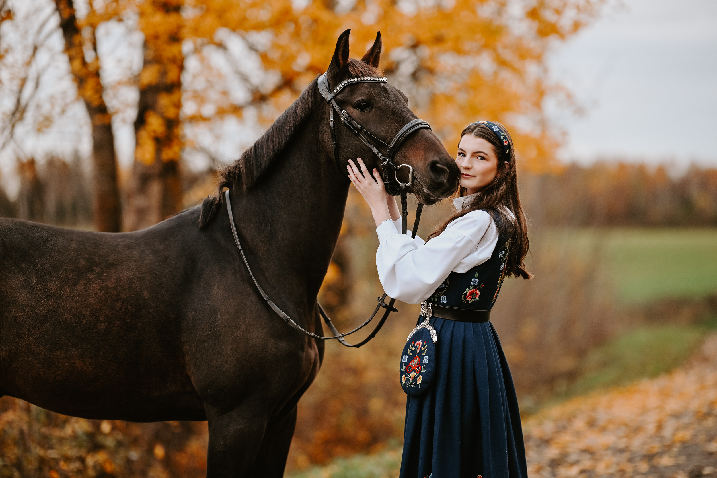 En kvinne i tradisjonell norsk bunad som står ved siden av en svart hest i høstlandskap.