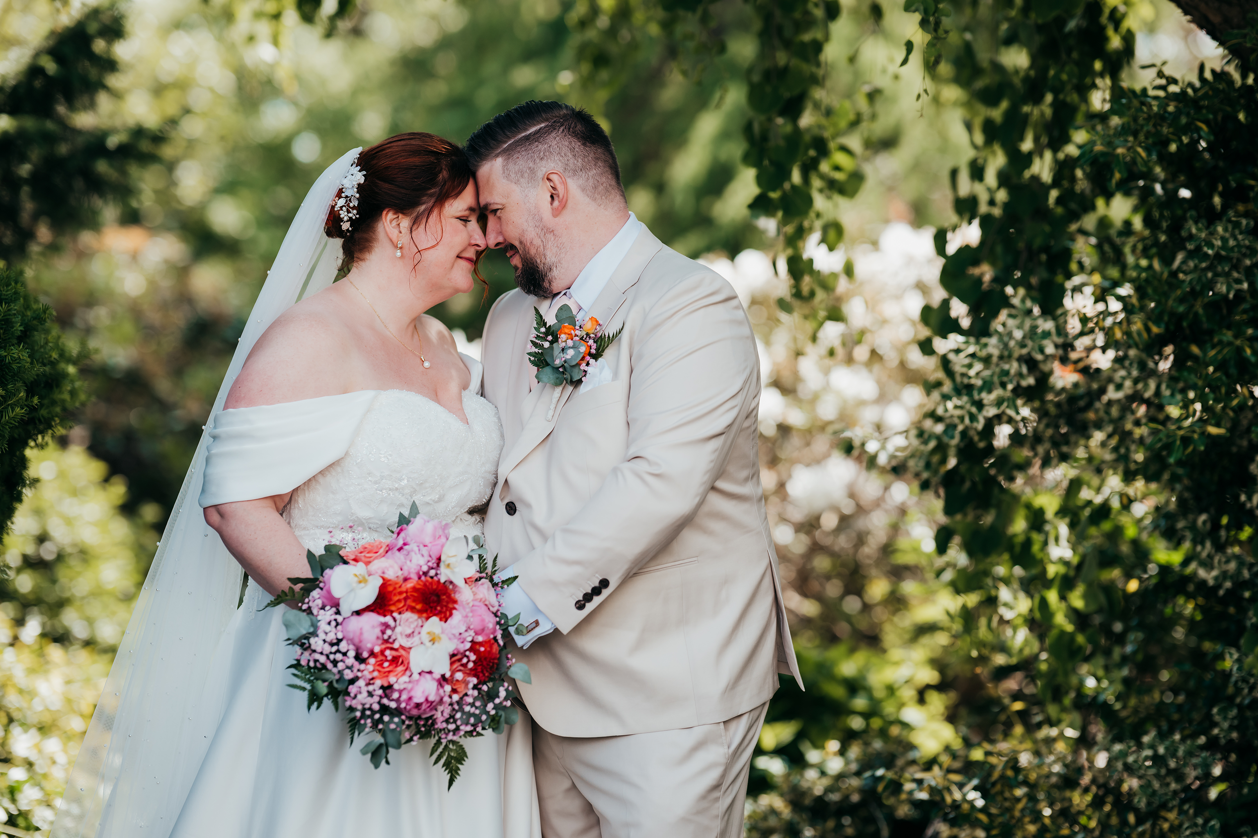 Brudepar står tett sammen, med pannen og nesen mot hverandre under en bånd av grønne løvverk, og smiler i kjærlighet. Bruden har på seg en hvit kjole med avskårne skuldre og holder en fargerik blomsterbukett. Brudgommen er i en lysebeige dress med blomsterlapp i kneget. Bakgrunnen er fylt med grønt løvverk i utendørs setting.
