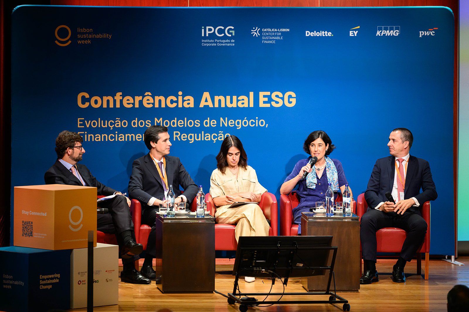 A panel of five people sitting on stage at a conference, with a blue backdrop displaying logos and the text 'Conferência Anual ESG' and elaborating on business models, financing, and regulation.