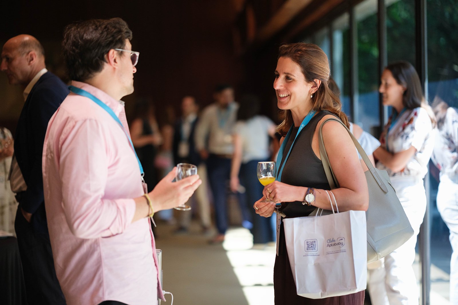 Two women are engaged in a conversation at a professional event, holding wine glasses, with others in the background.