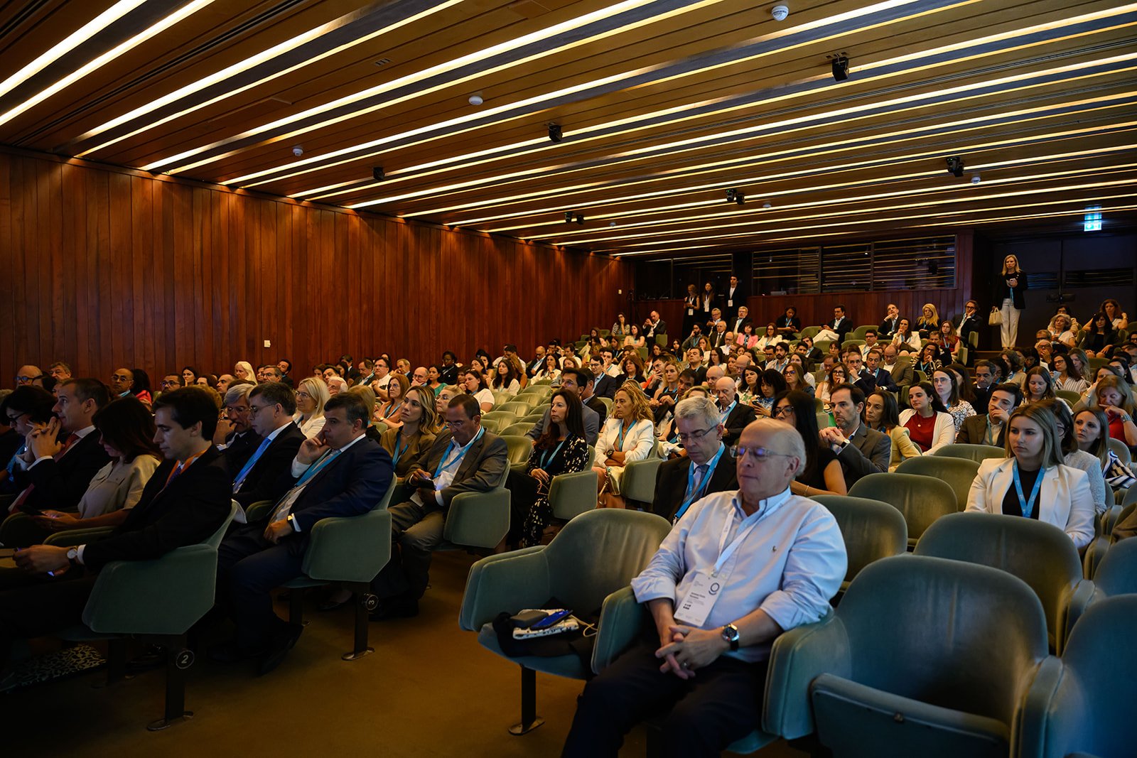 Large audience seated in a conference hall with wooden walls and ceiling, attending a formal event.