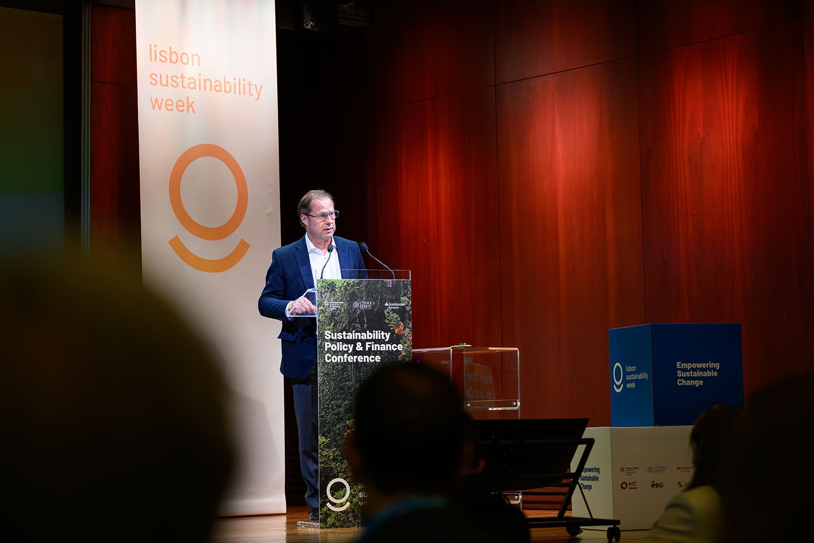 Man giving a presentation at the Sustainability Policy & Finance Conference during Lisbon Sustainability Week, standing behind a podium with a banner and a screen in the background.