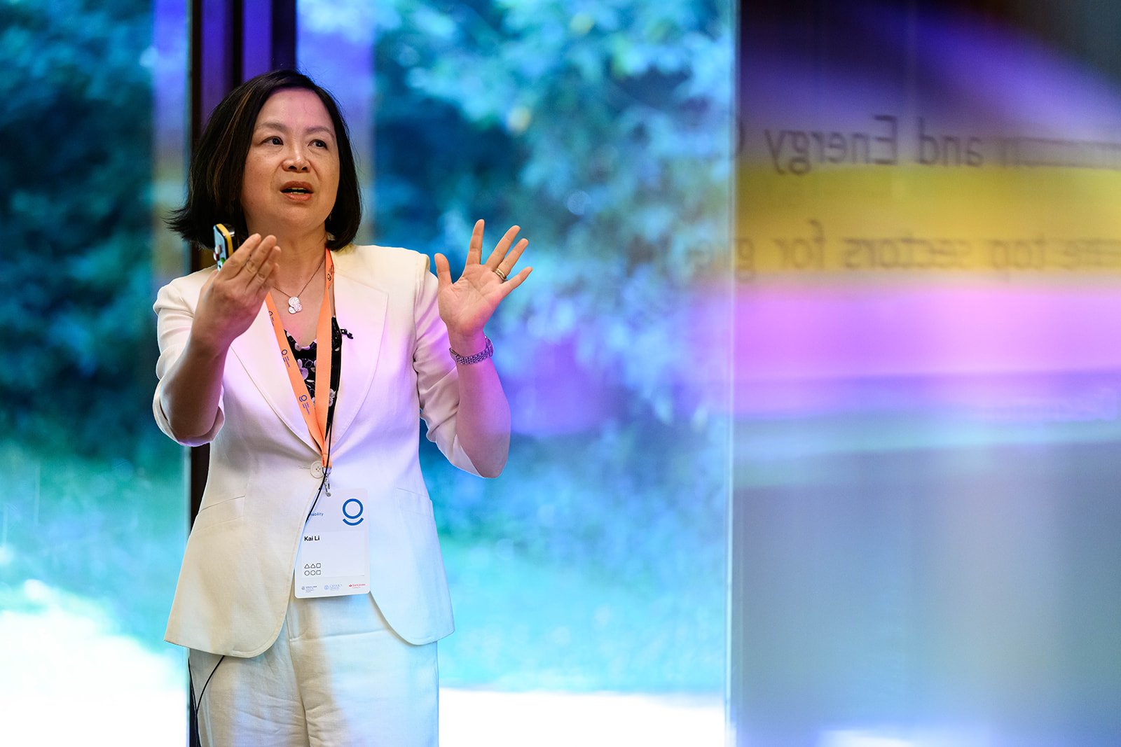 A woman wearing a white suit and an orange lanyard is giving a presentation or speech in front of a colorful background.