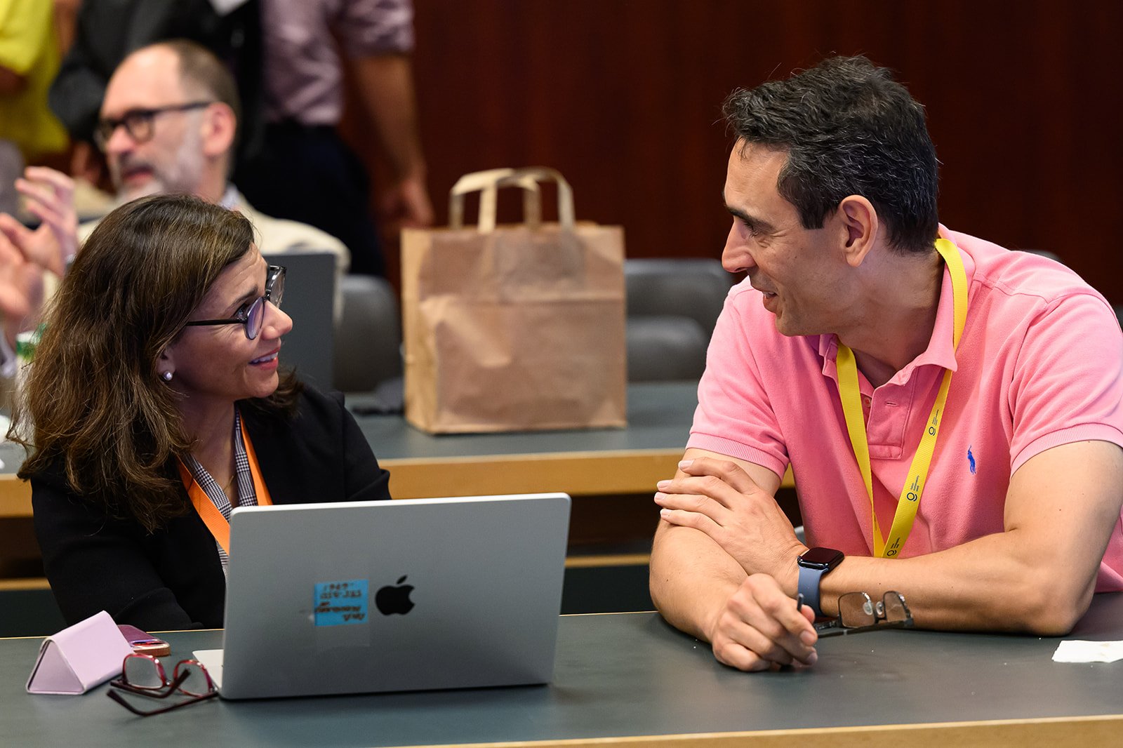 A woman and a man engaging in conversation at a conference or meeting. The woman has shoulder-length brown hair, glasses, and is wearing a black jacket. The man has short dark hair, a pink polo shirt, and a yellow lanyard. They are sitting at a table