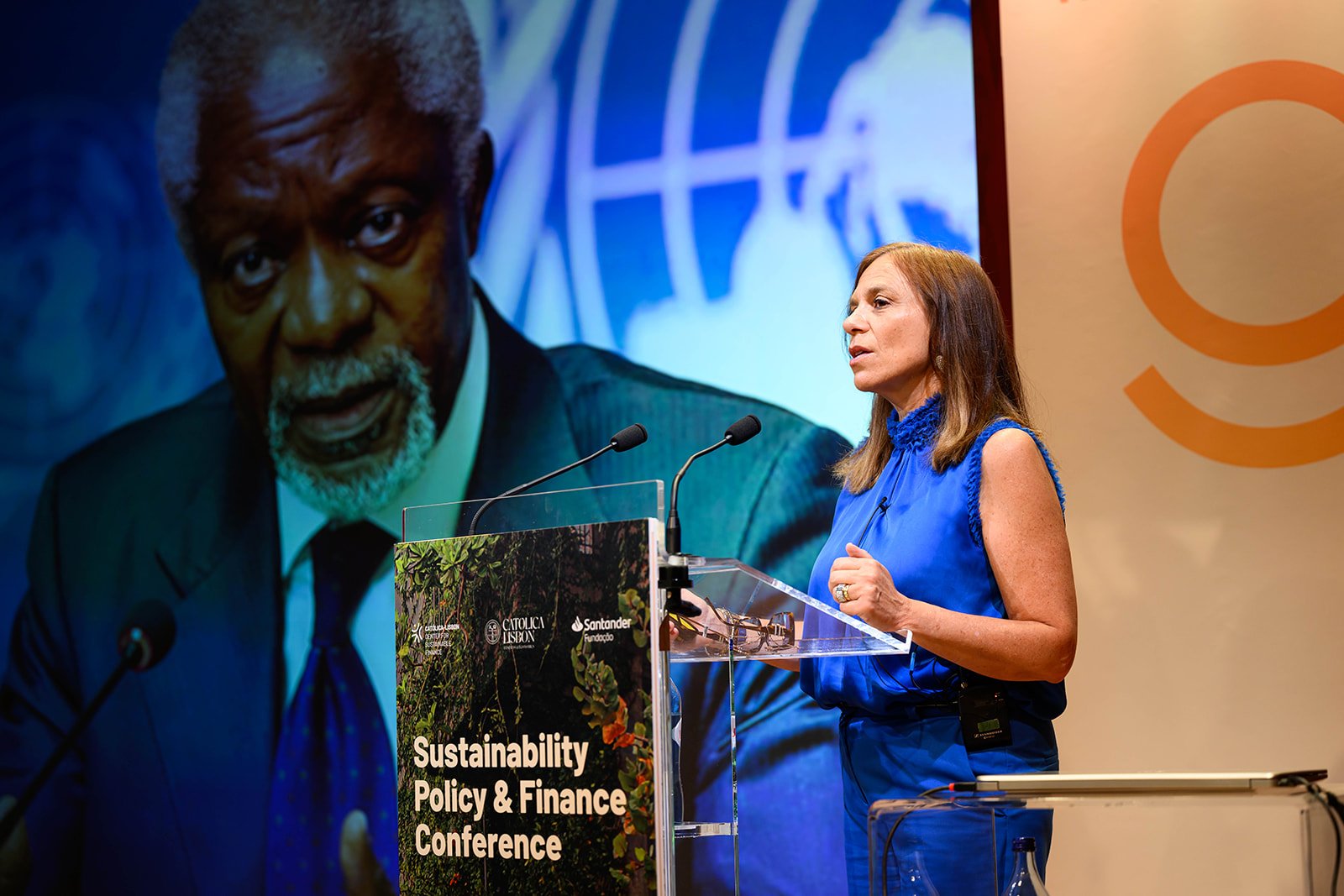 A woman in a blue dress speaks at a Sustainability Policy & Finance Conference. She stands at a podium with a microphone, with a large screen behind her displaying an older man in a suit and tie.