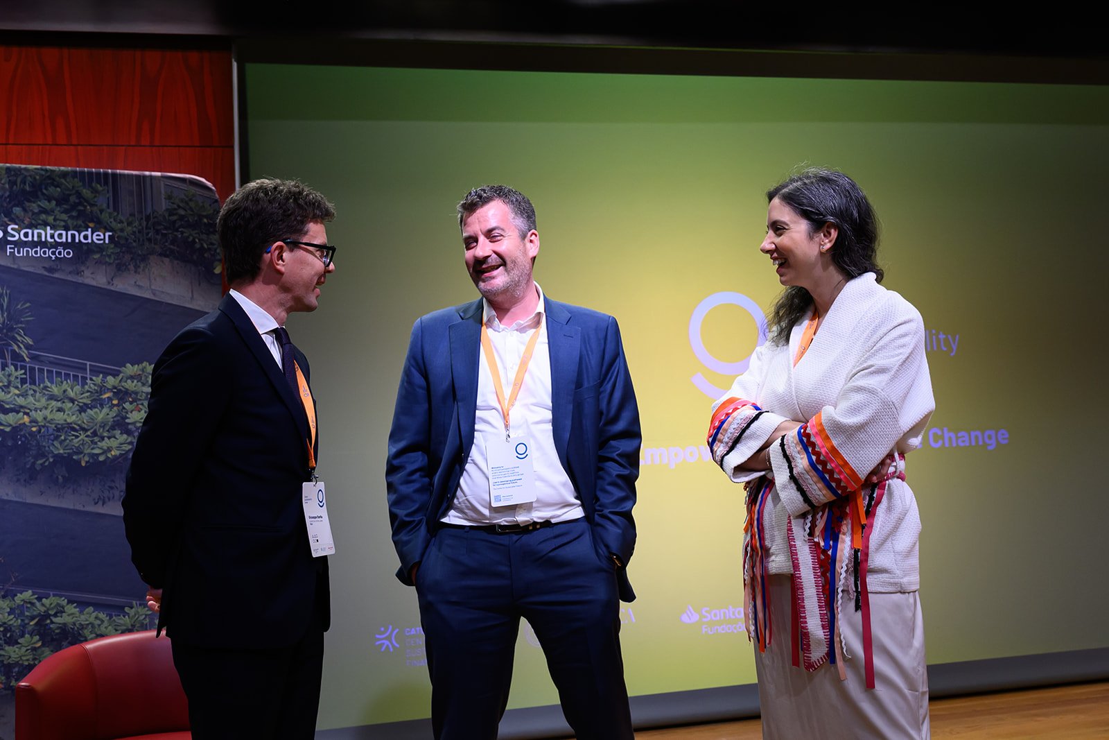 Three people in formal attire having a conversation on stage at an event, with a backdrop displaying the logos of Santander and a yellow and purple design.