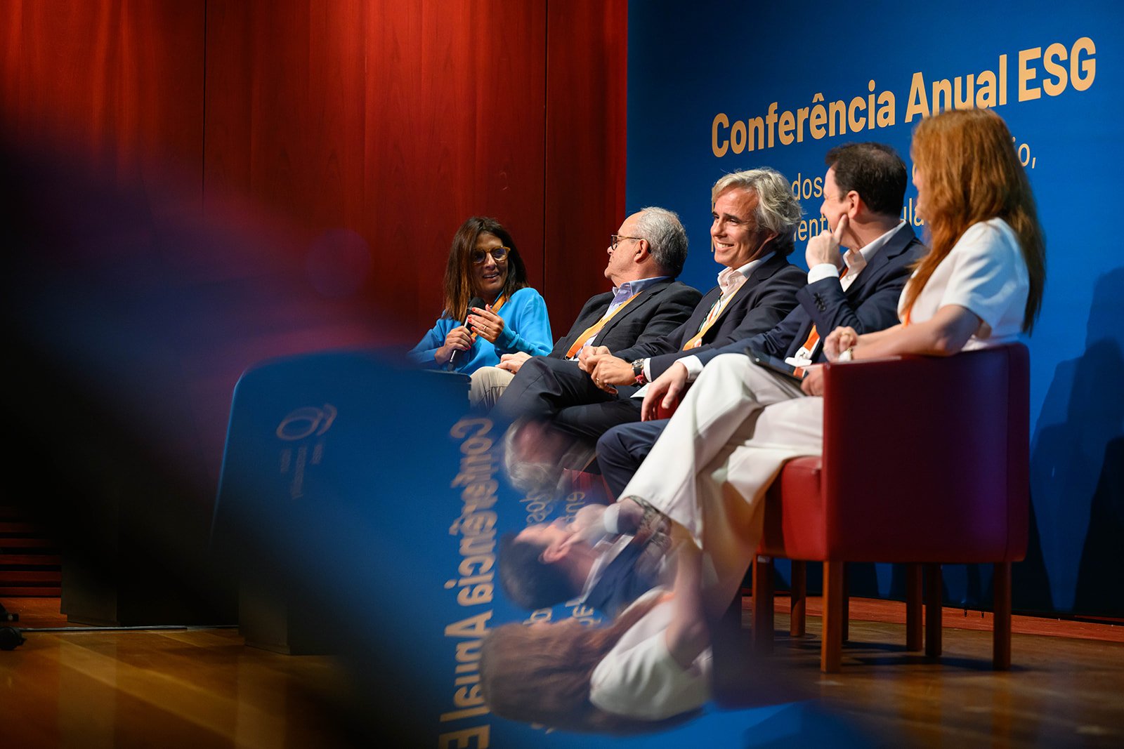 Panel of six people seated on a stage during a conference, with a blue backdrop displaying text in Portuguese. One woman is speaking into a microphone, and the others are listening attentively.