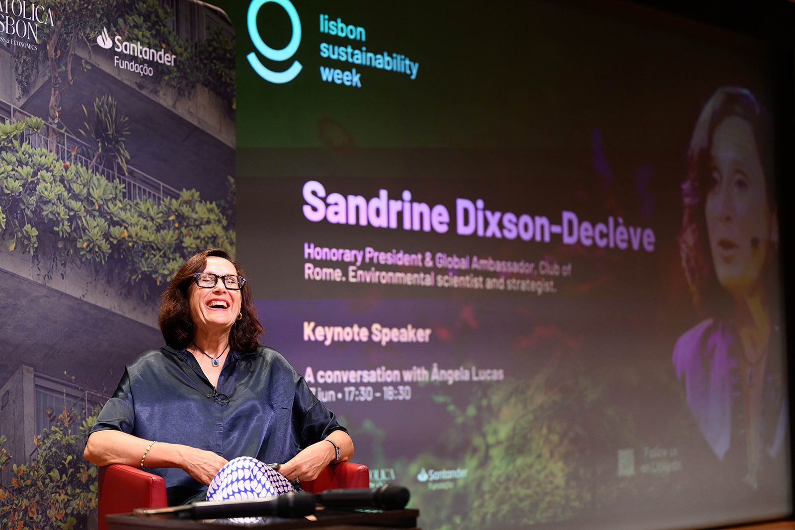 A woman with glasses and dark hair, wearing a black blouse, laughing while sitting on a red chair in front of a large screen at an event. The screen displays a presentation titled 'Lisbon Sustainability Week' with the name Sandrine Dixson-Declève and