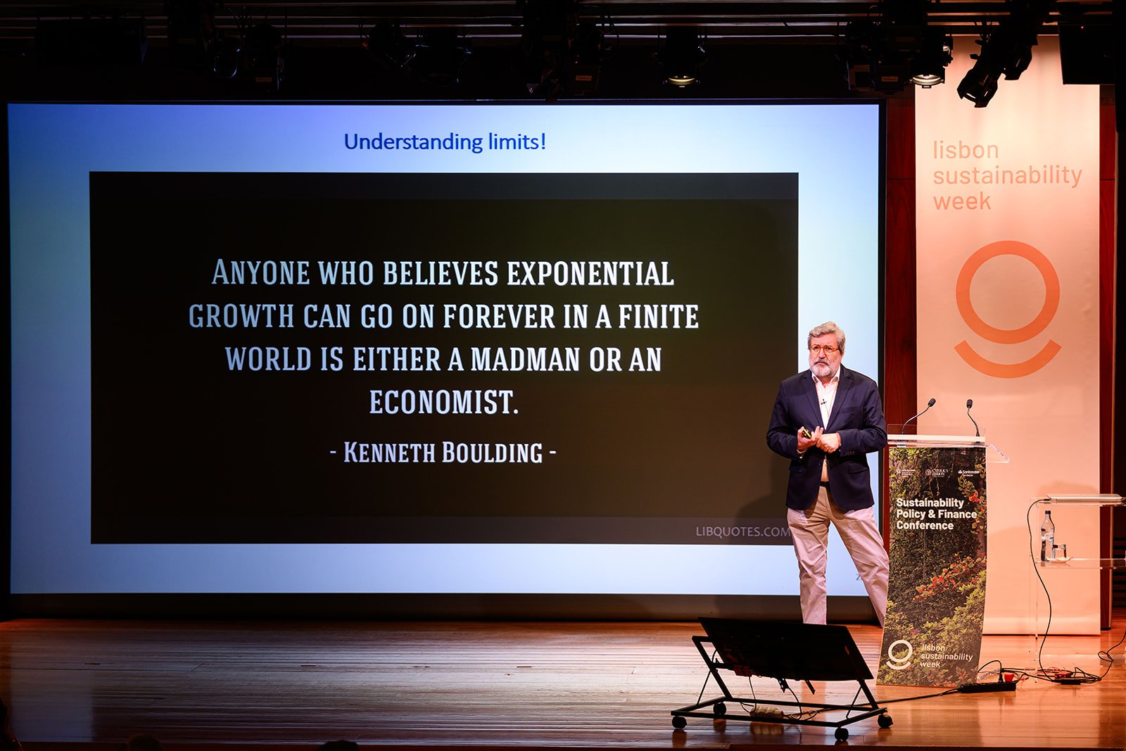 Male speaker gives presentation at Lisbon Sustainability Week standing next to a large screen displaying a quote about exponential growth, with a podium labeled 'Sustainability Policy & Finance Conference' and a backdrop with 'lisbon sustainability w