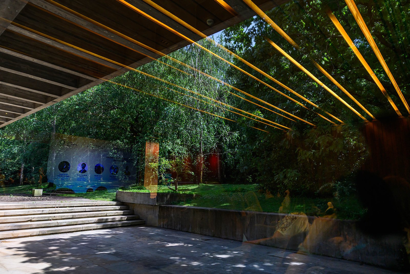 A modern building with a large glass window reflecting trees and a park outside, featuring yellow light strips on the ceiling and concrete stairs leading out.