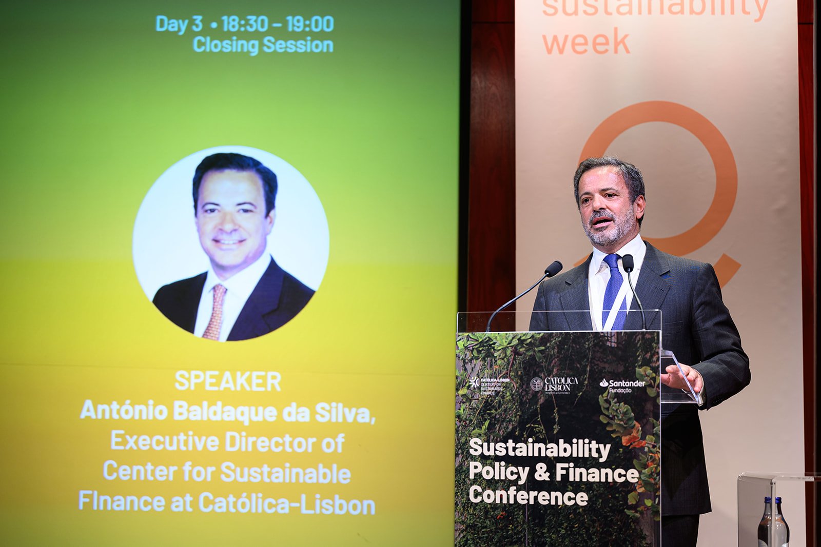Man in a dark suit and blue tie speaking at a podium during a sustainability conference, with a large screen displaying his name and photo, and a banner that reads 'Sustainability Week' and 'Sustainability Policy & Finance Conference' in the backgrou