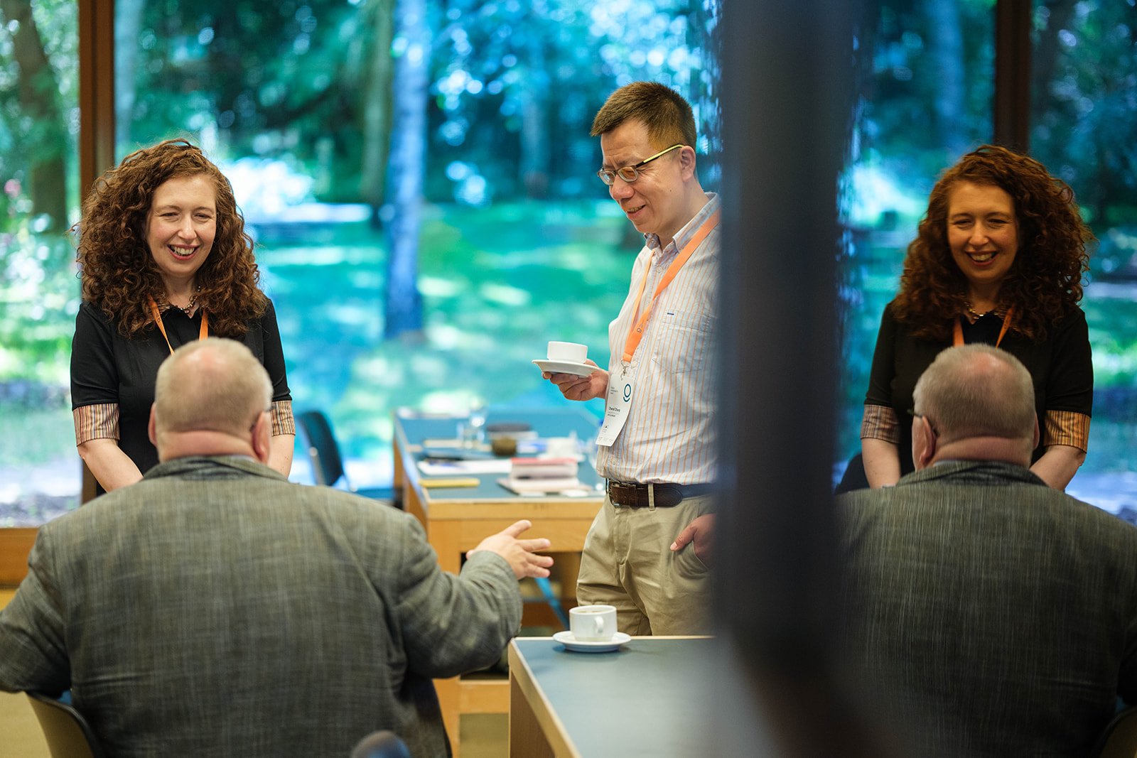 Two women and a man are standing and conversing with two men seated at a table during a professional event. The women are smiling, and the man standing is holding a cup and saucer, also smiling. The background shows a large window with a view of tree