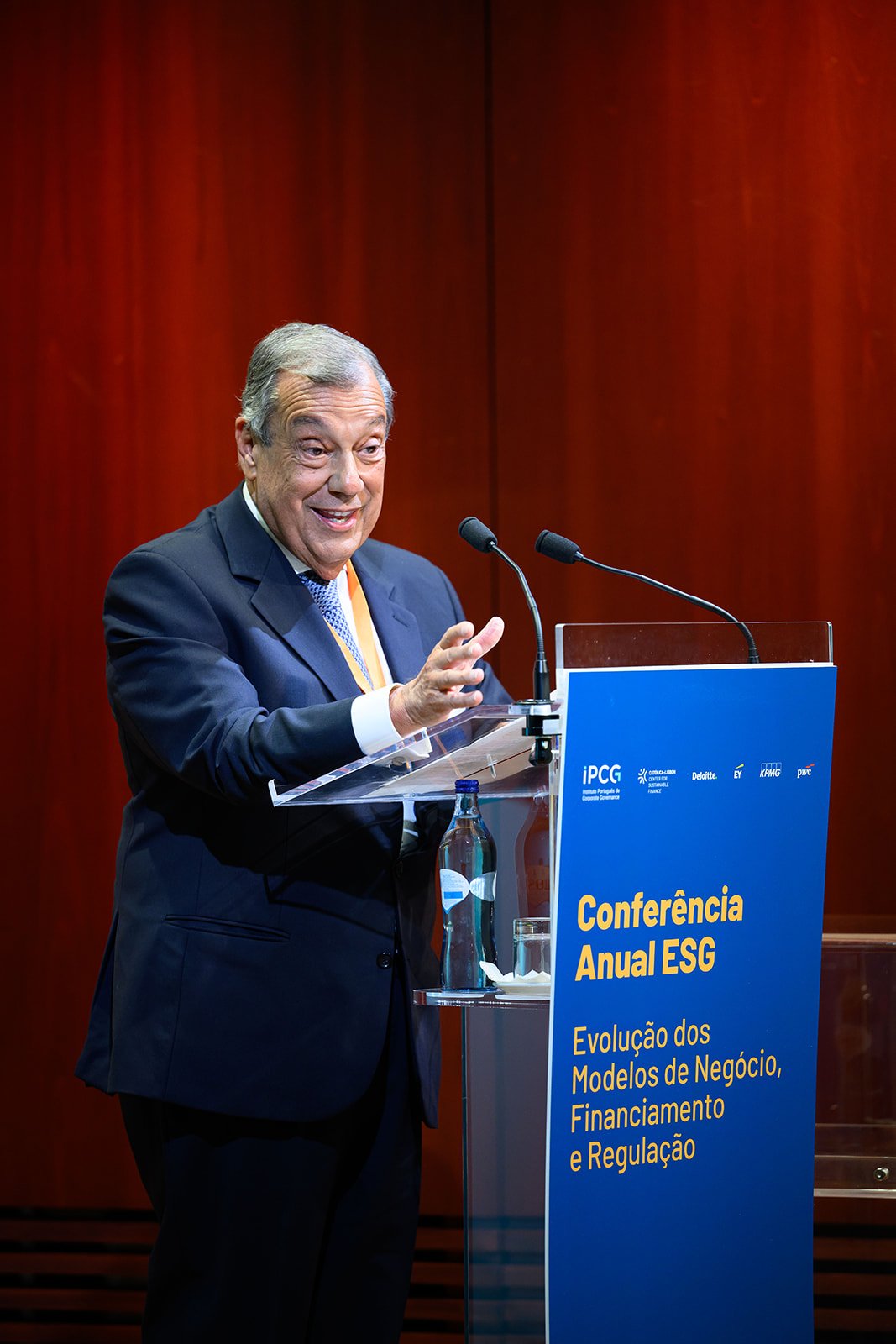 A man in a dark suit speaking at a podium with a blue sign, at an annual ESG conference, with a redwood wall background.