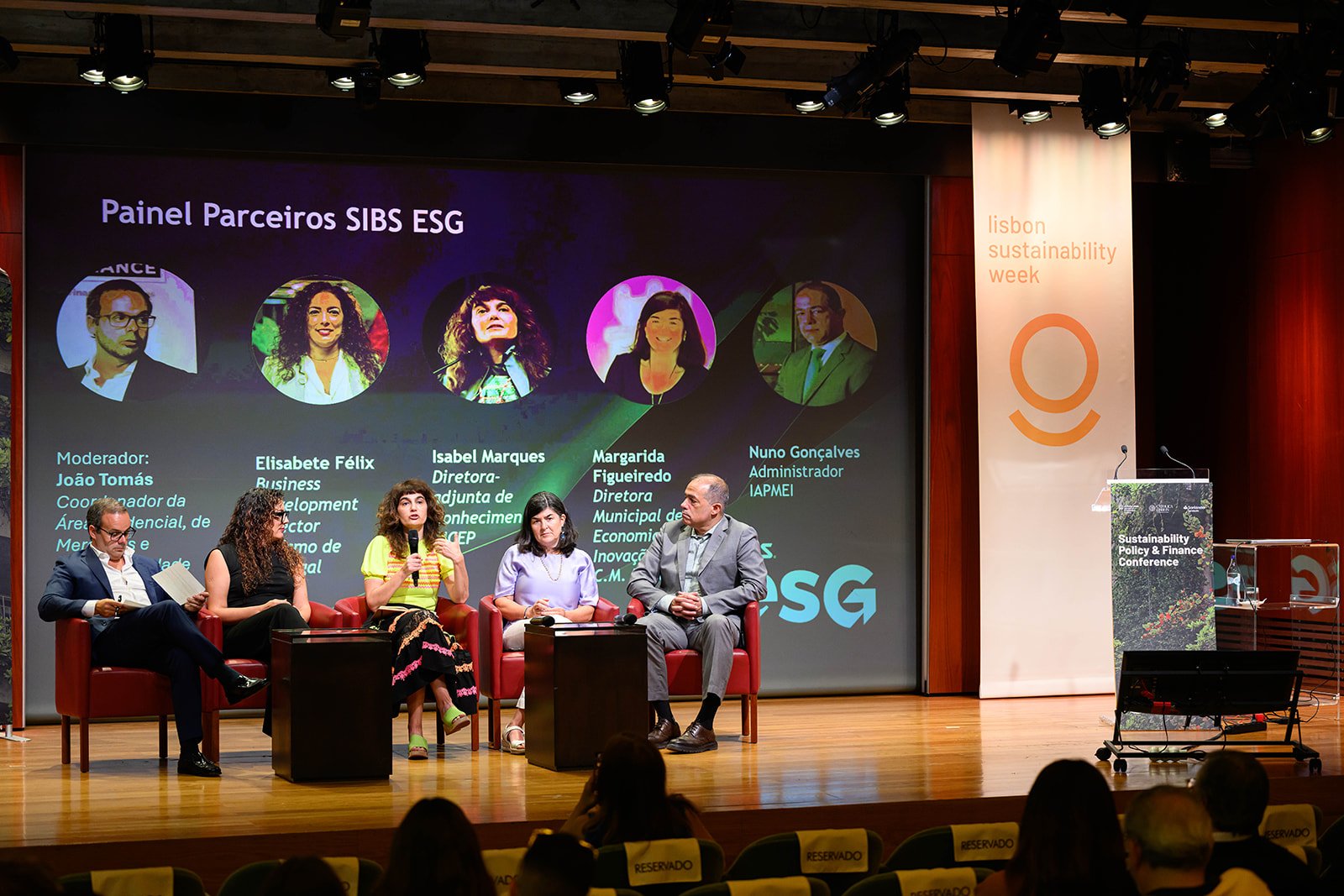 A panel discussion at the Lisbon Sustainability Week with five speakers seated on stage, and a large screen behind them displaying the panelists' names and roles. The background of the screen shows a title in Portuguese and portraits of the panelists