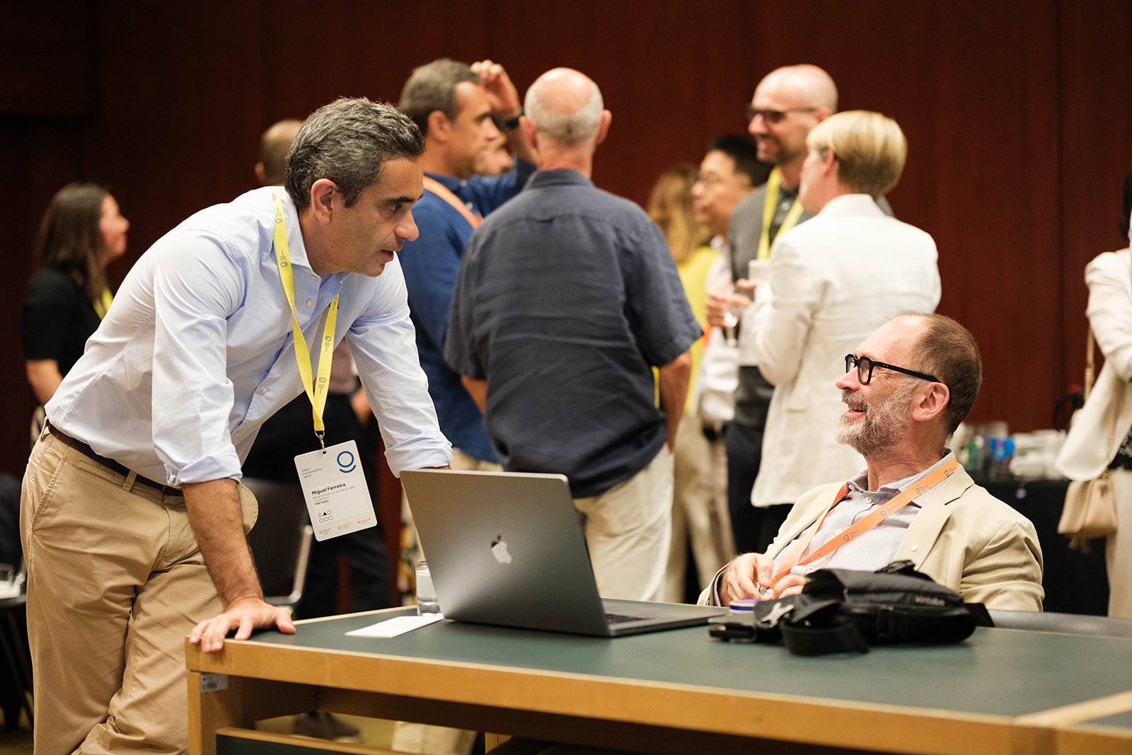 Two men having a conversation at a conference, one standing and leaning on a table, the other sitting with a laptop, in a room full of people networking.