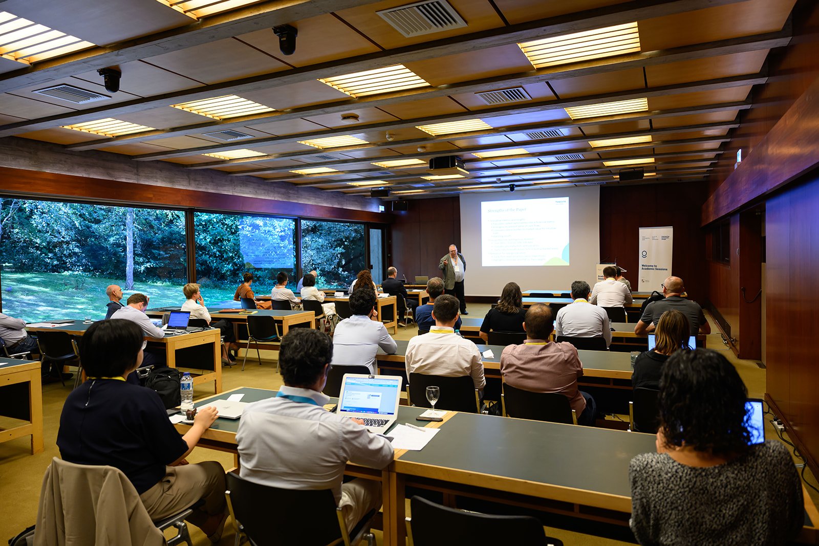 A man giving a presentation to an audience in a conference room with large windows showing trees outside. The room has a screen displaying slides and several people seated at tables using laptops and taking notes.