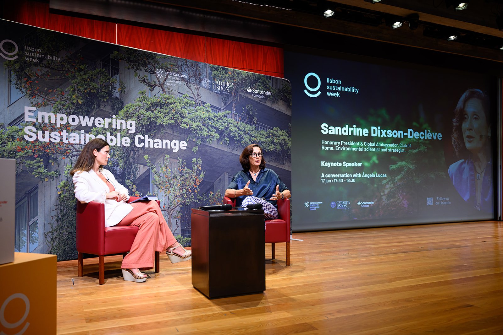 Two women are seated on stage at Lisbon Sustainability Week. One woman, on the right, is speaking and wearing glasses, a dark blue top, and patterned pants. The other woman, on the left, is listening and wearing a white blazer and peach pants. There