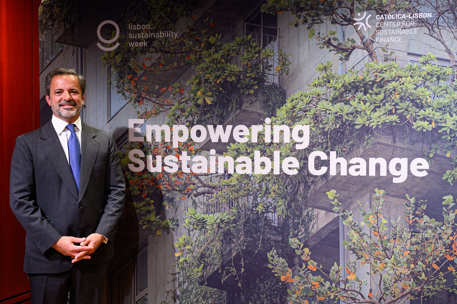 A man in a dark suit standing next to a large banner with the text "Empowering Sustainable Change" at an event related to Lisbon Sustainability Week. The banner features greenery and a building in the background.