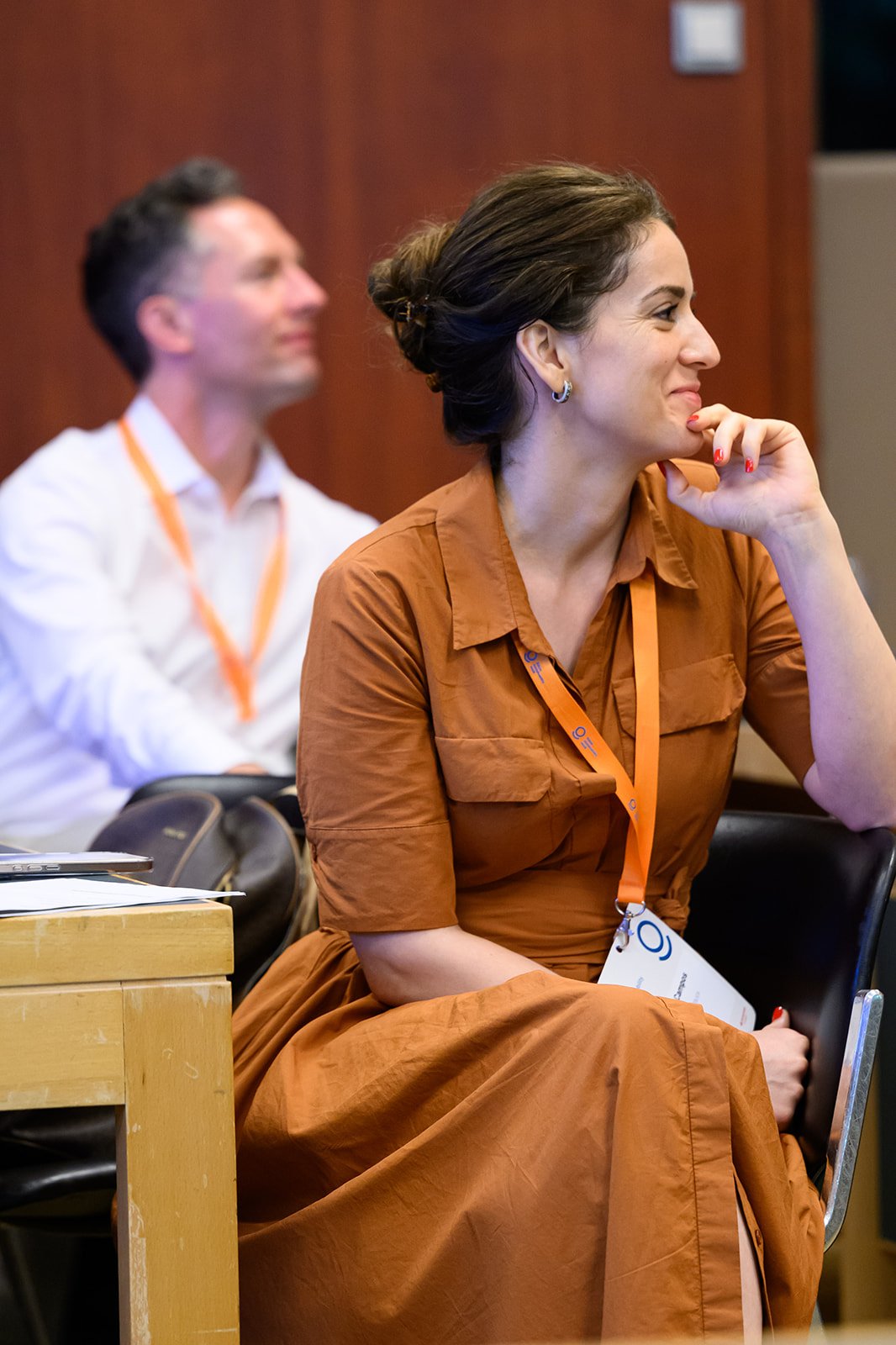 A woman sitting in a conference room, smiling and wearing a brown dress with an orange badge around her neck.