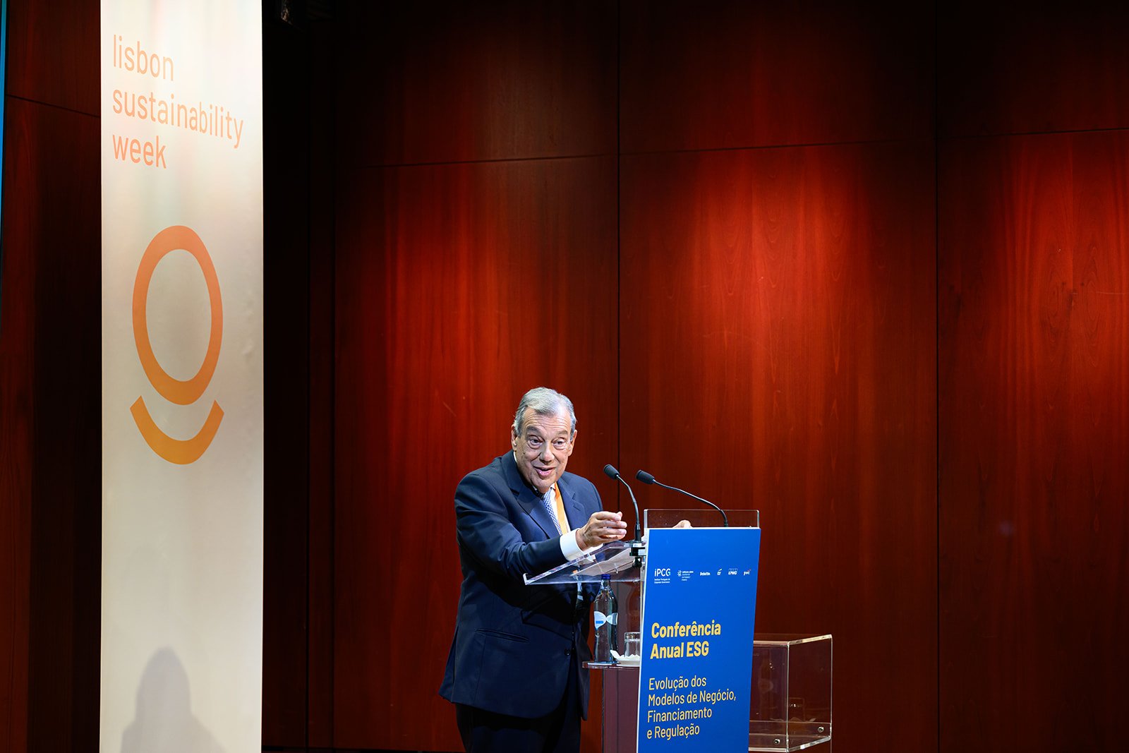 A man in a suit speaking at a podium during a conference with a sign that reads 'Lisbon Sustainability Week' and 'Conferência Anual ESG'. The background features wooden paneling.