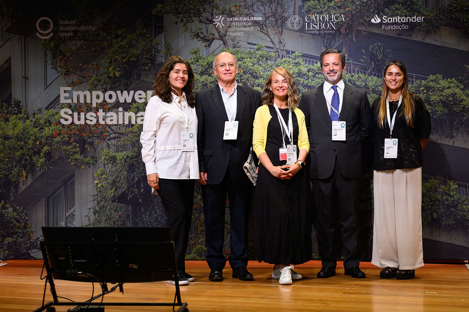 Group of six people posing on stage during Lisbon Sustainability Week event, with a backdrop displaying green foliage and logos of sponsors, including Católica Lisbon and Santander.
