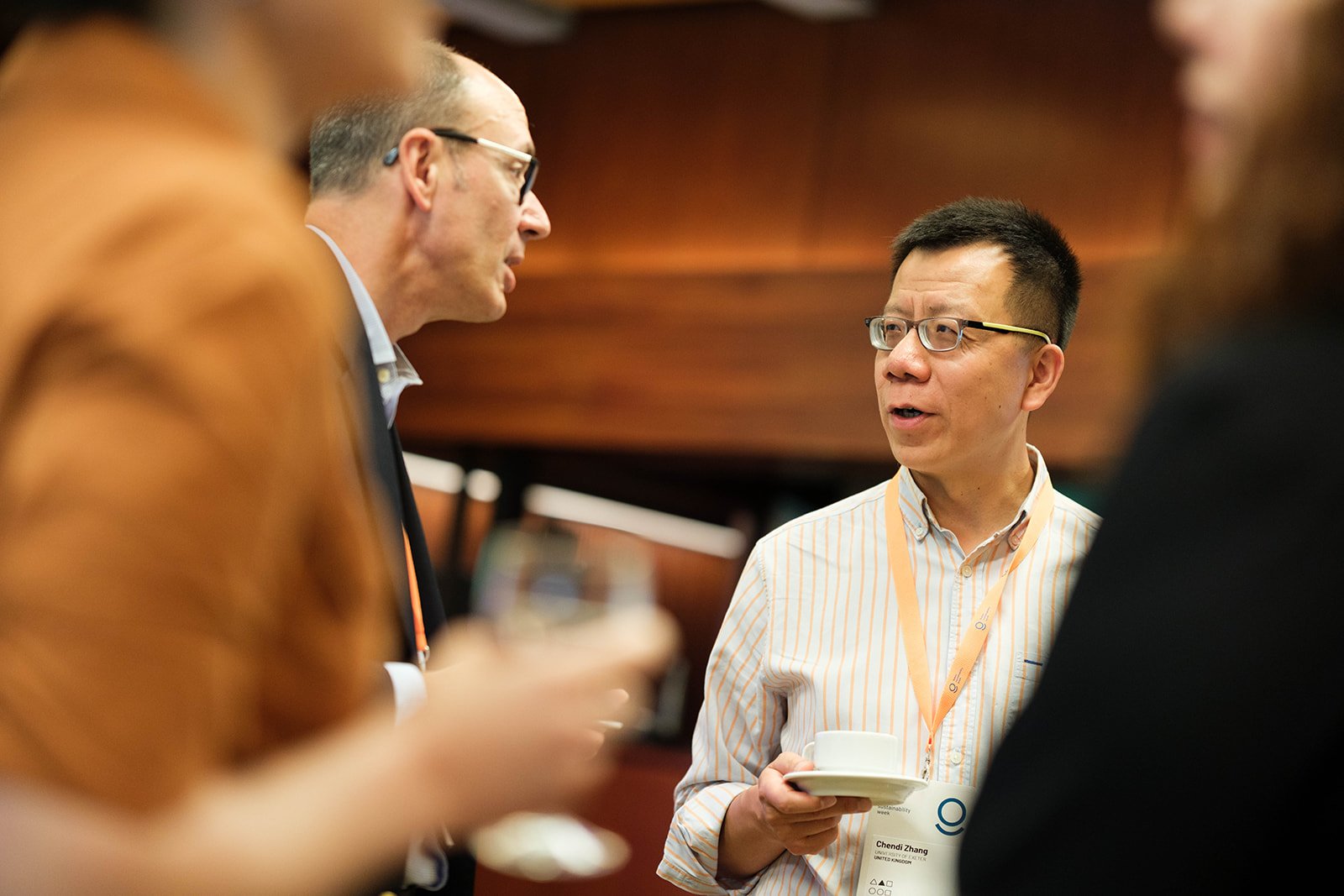A group of professionals having a conversation at a conference, one person holding a coffee cup and plate, with name tags visible.