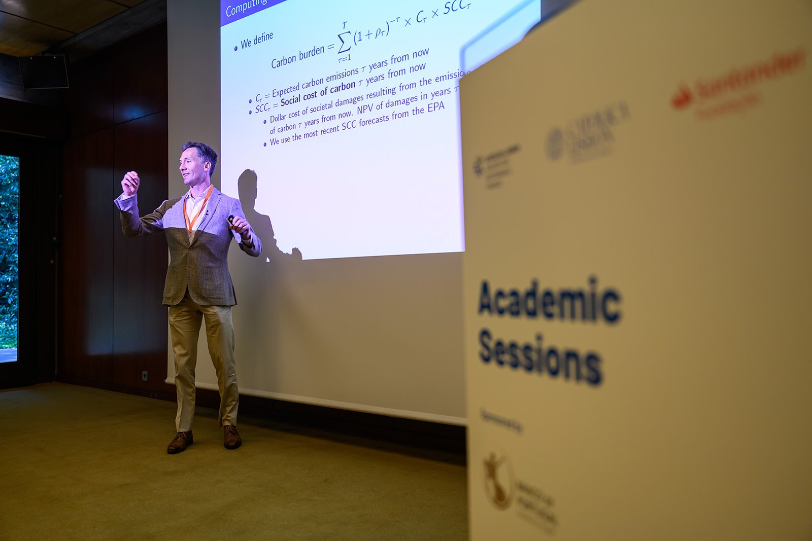 A man in a brown and beige suit and khakis giving a presentation at an academic conference, with a slide on carbon burden projected on the screen behind him, and a yellow sign in the foreground reading 'Academic Sessions'.