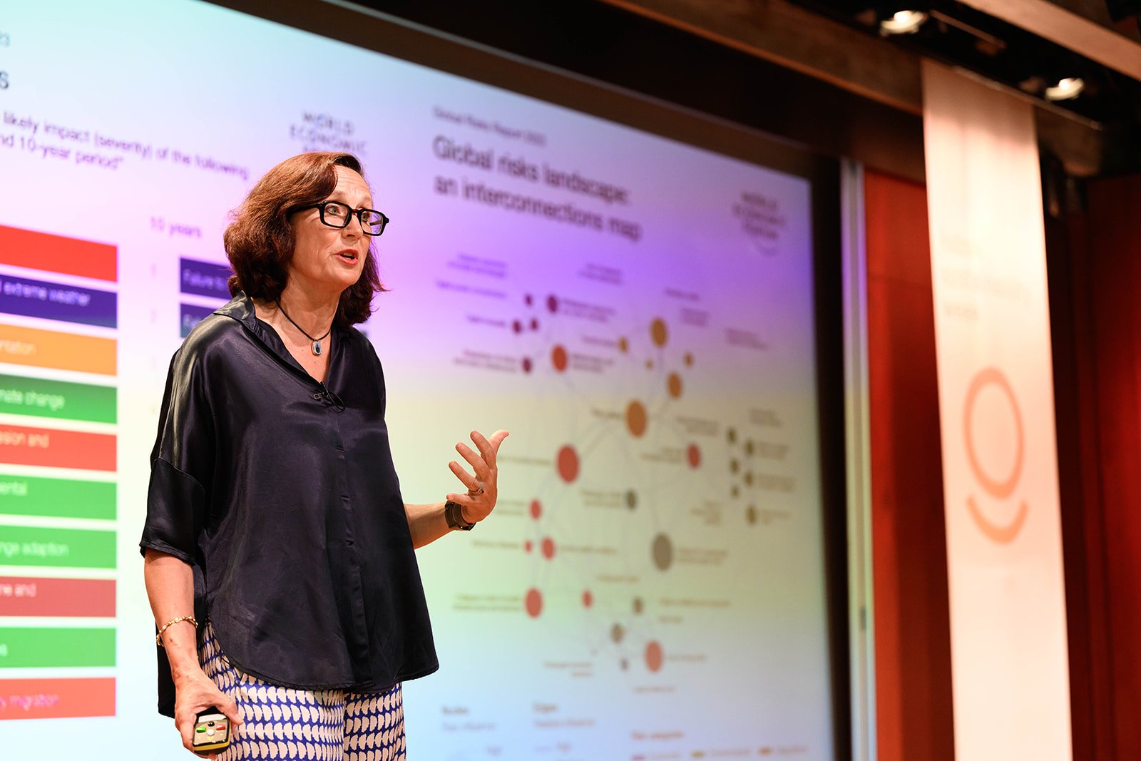 A woman with dark brown hair, glasses, and a black satin shirt giving a presentation in front of a large colorful infographic on a screen.