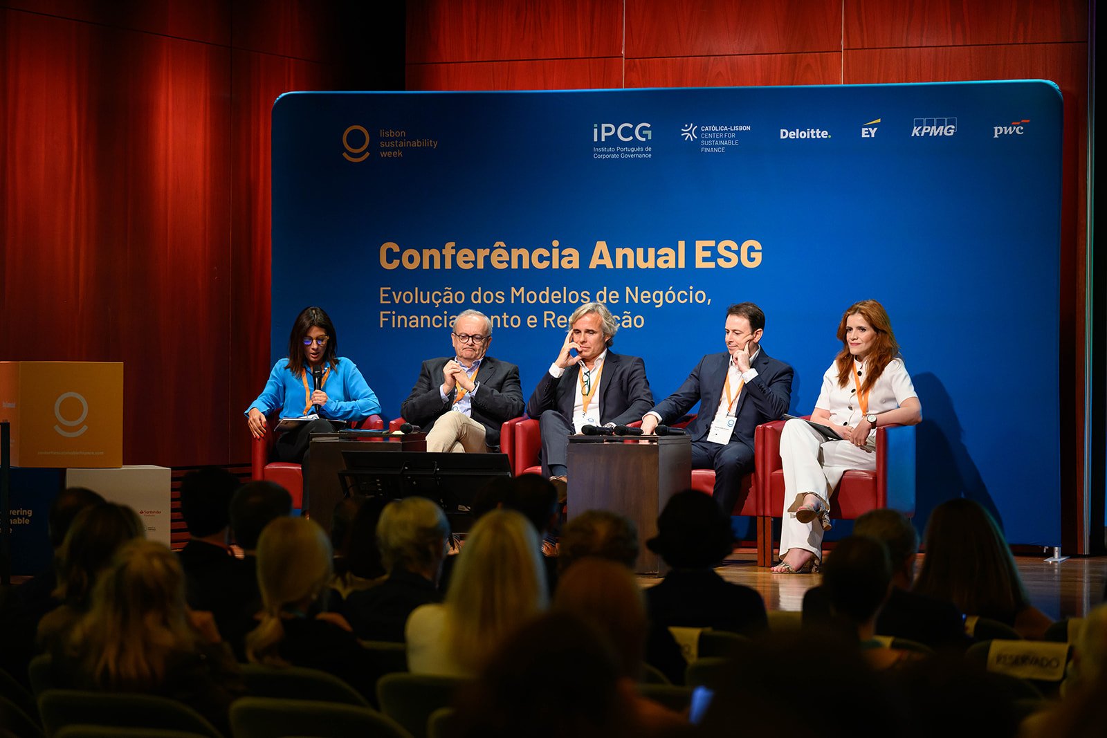 Panel of five speakers seated on stage at the Lisbon Sustainability Week conference. The background displays a blue banner with the conference title, sponsors, and logos. Audience members seated in front are visible.
