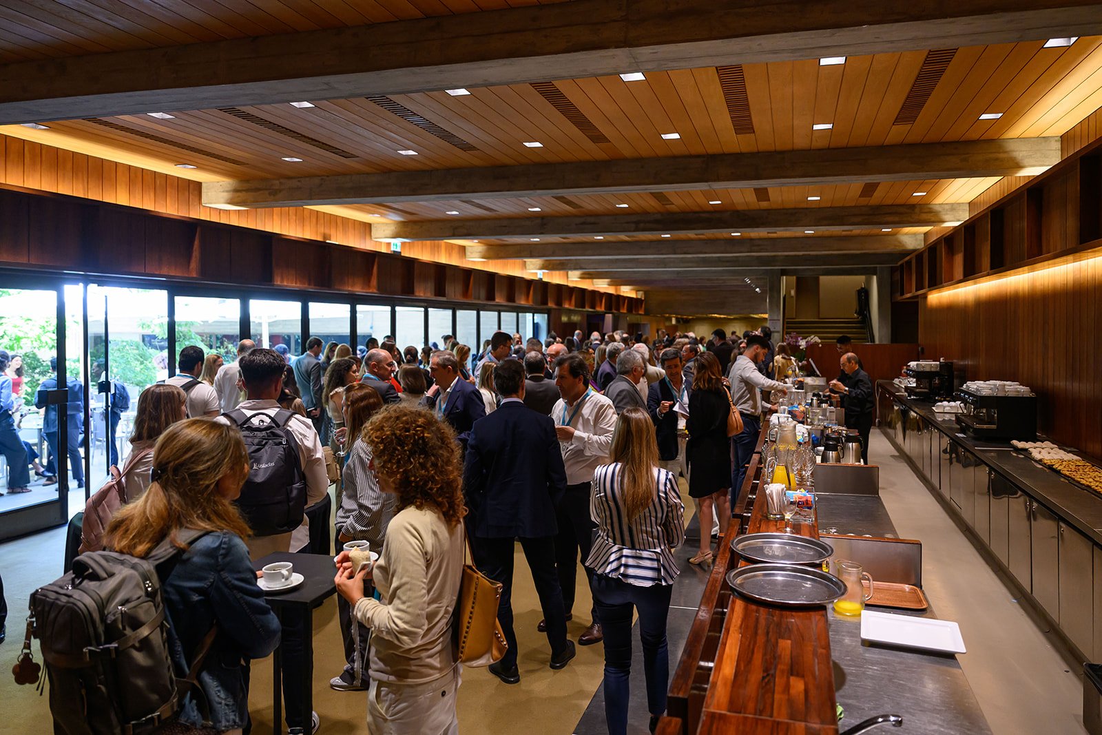 Crowd of people at a hotel or conference buffet in a modern, wood-paneled interior with large windows.