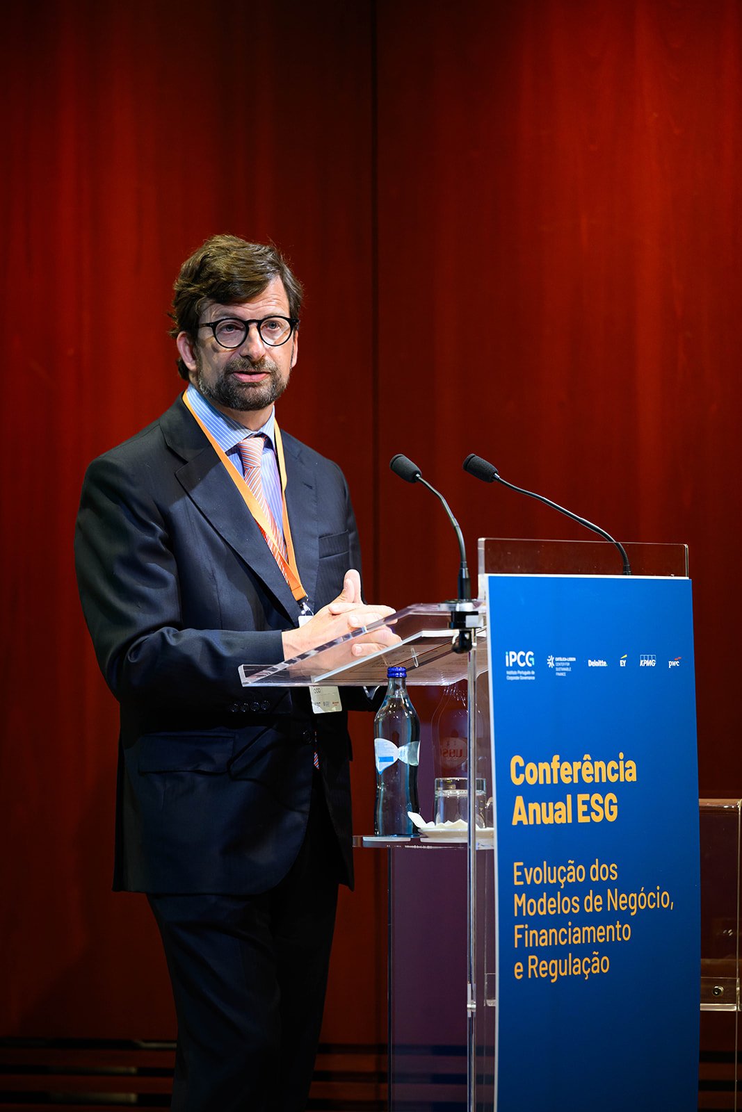 A man in a dark suit and glasses standing at a podium with a blue sign at a conference, with a red wooden wall in the background.