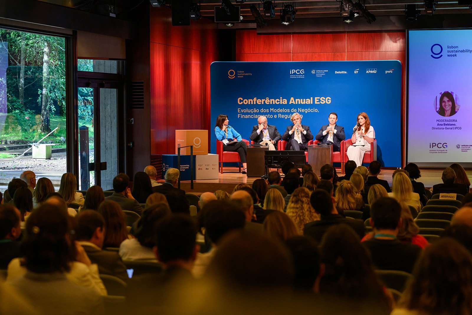 A panel discussion at the Lisbon Sustainability Week conference with five speakers on stage, a large blue banner behind them, and a seated audience watching inside a conference hall.