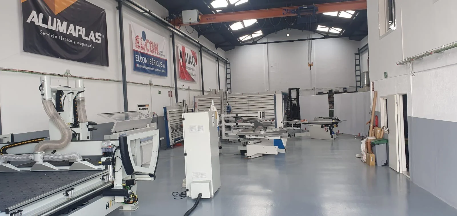 Interior de un taller o fábrica con varias máquinas de corte y ensamblaje, paredes blancas, banners publicitarios, y un techo con vigas metálicas y cristaleras, en un entorno ordenado y bien iluminado.