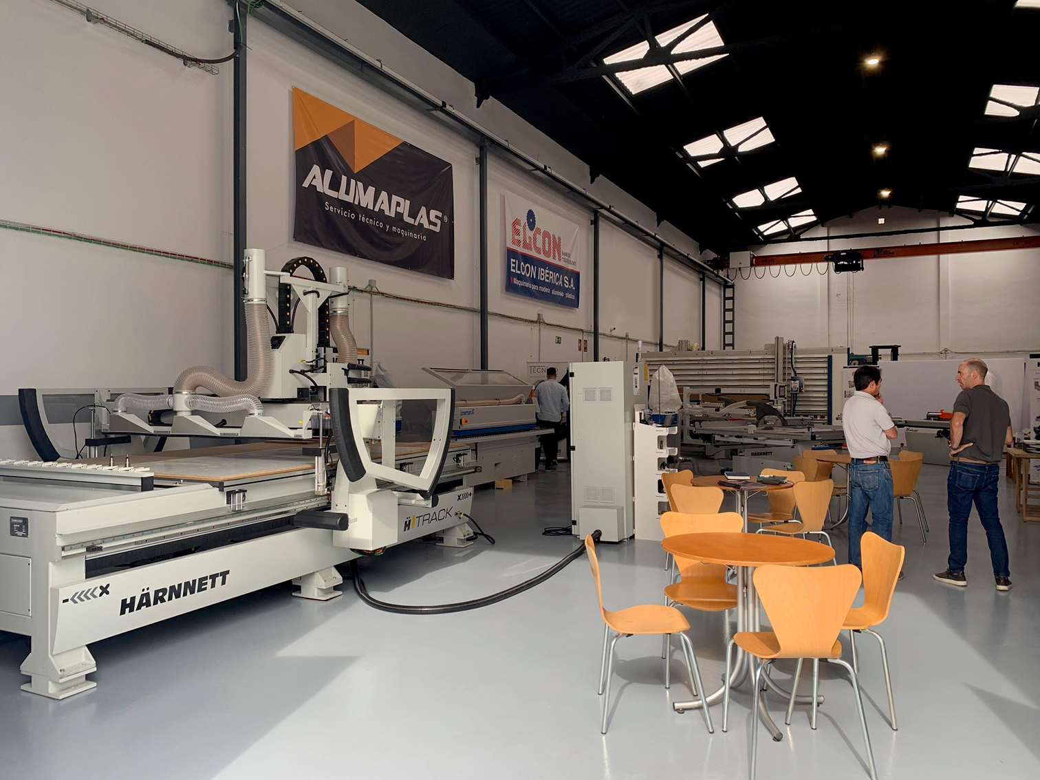 Interior de una fábrica o taller con maquinaria industrial y dos personas conversando, y varias mesas y sillas en primer plano.