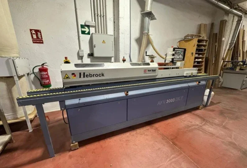 Una máquina de corte de madera marca Hebrock en un taller con madera apilada al fondo.