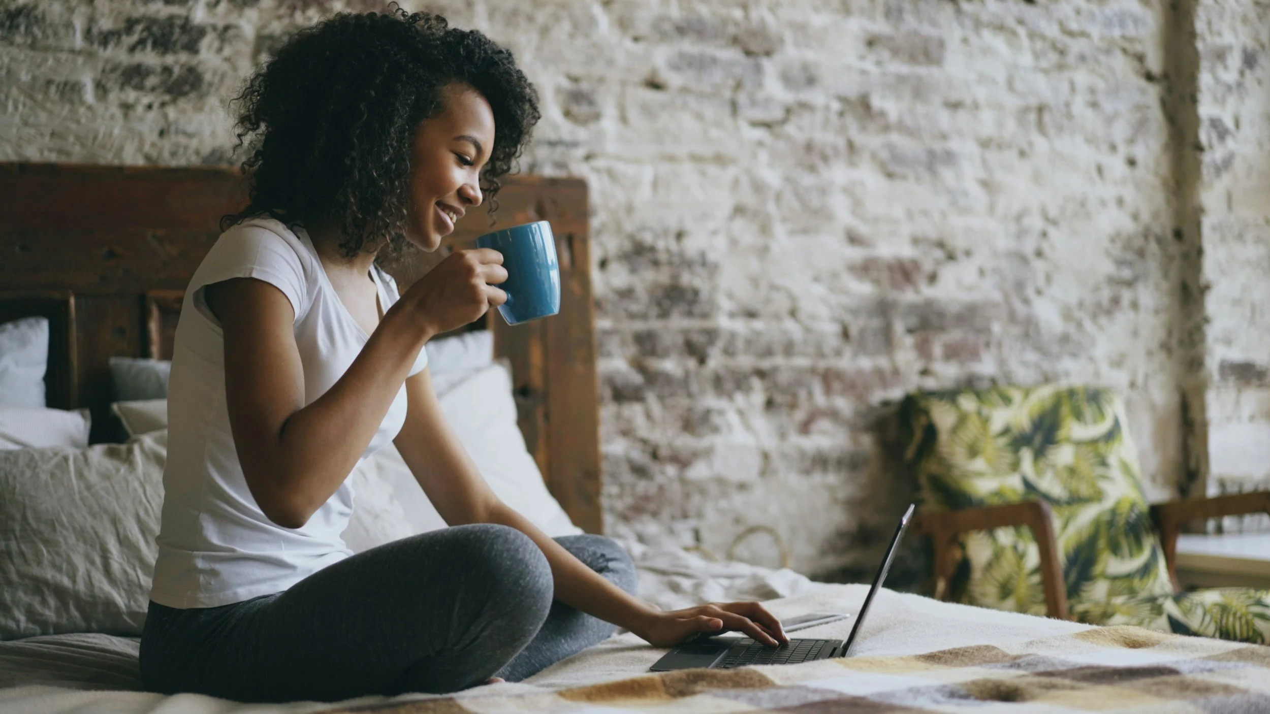 A woman sitting on a bed, smiling, and holding a blue mug, with a laptop on her lap in a cozy room with an exposed brick wall and decorative pillows.